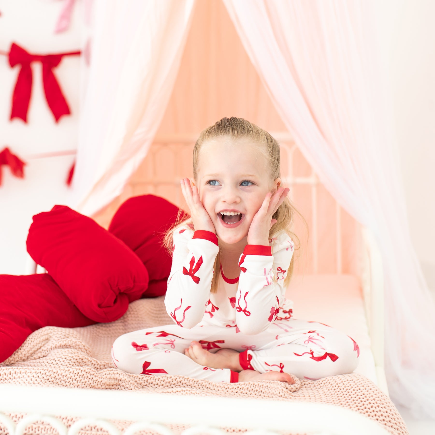 Young girl sitting on a bed wearing the Long Sleeve Pajamas in Small Love Bow with both her hands on her face