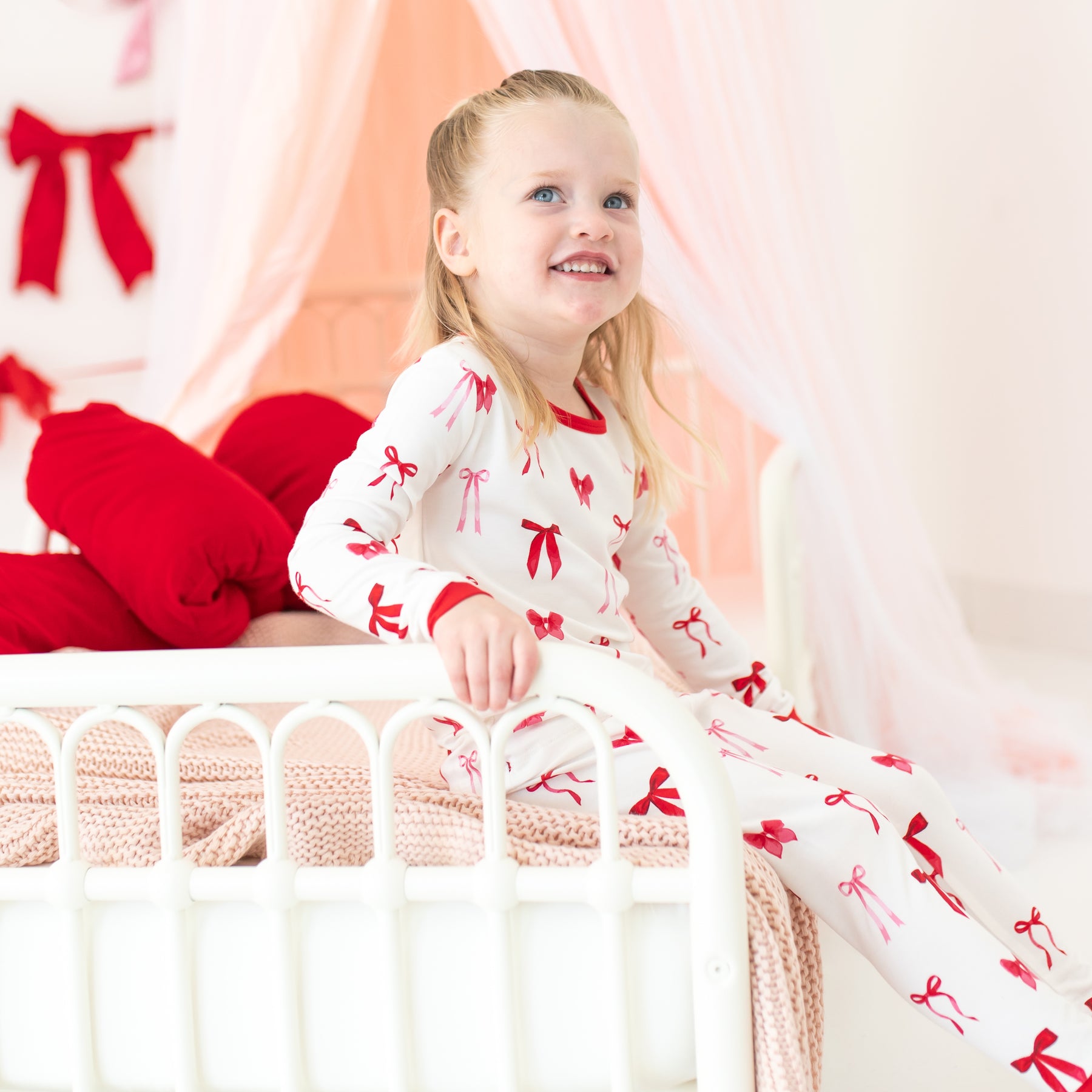 Young girl sitting on a bed wearing the Long Sleeve Pajamas in Small Love Bow