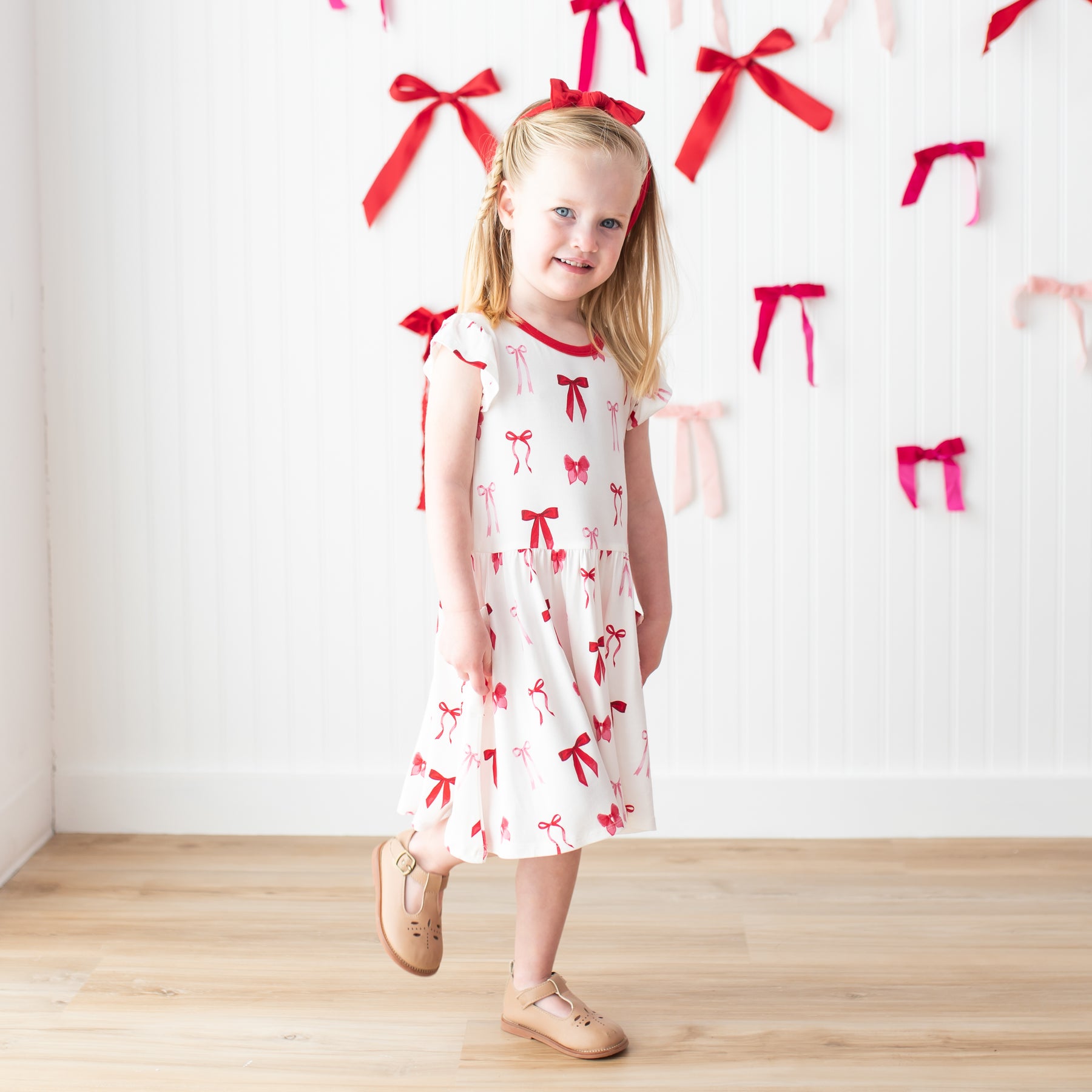 Young girl posing in front of a white paneled wall with bows on it wearing the Pocket Dress in Small Love Bow