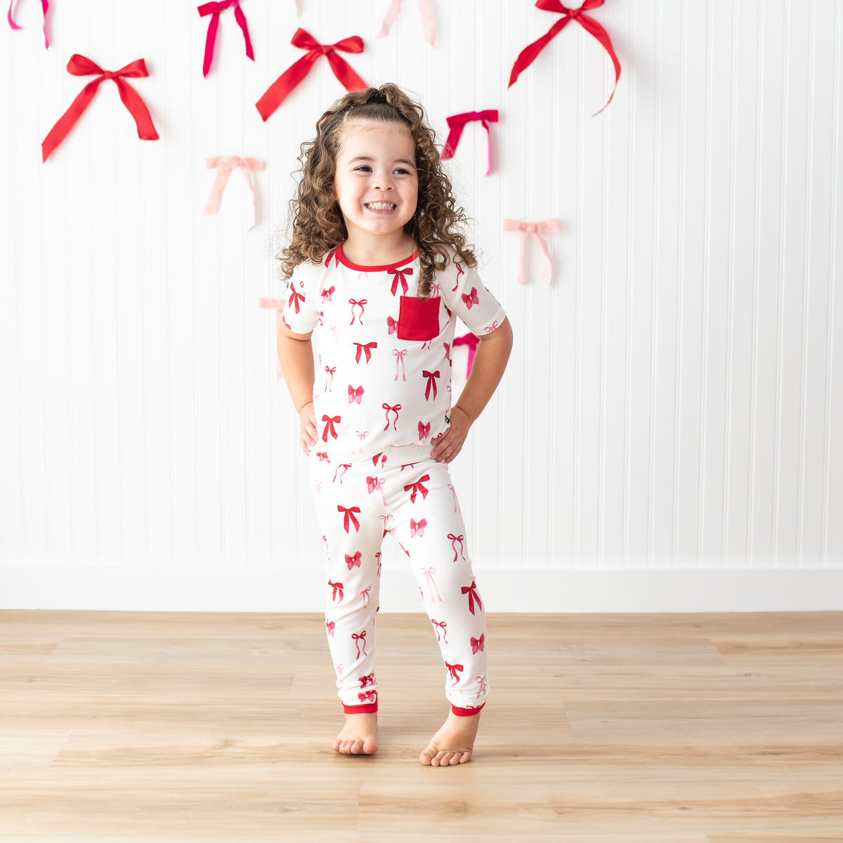 Young girl posing wearing the Short Sleeve with Pants Pajamas in Small Love Bow in front of a white paneled wall with bows on it