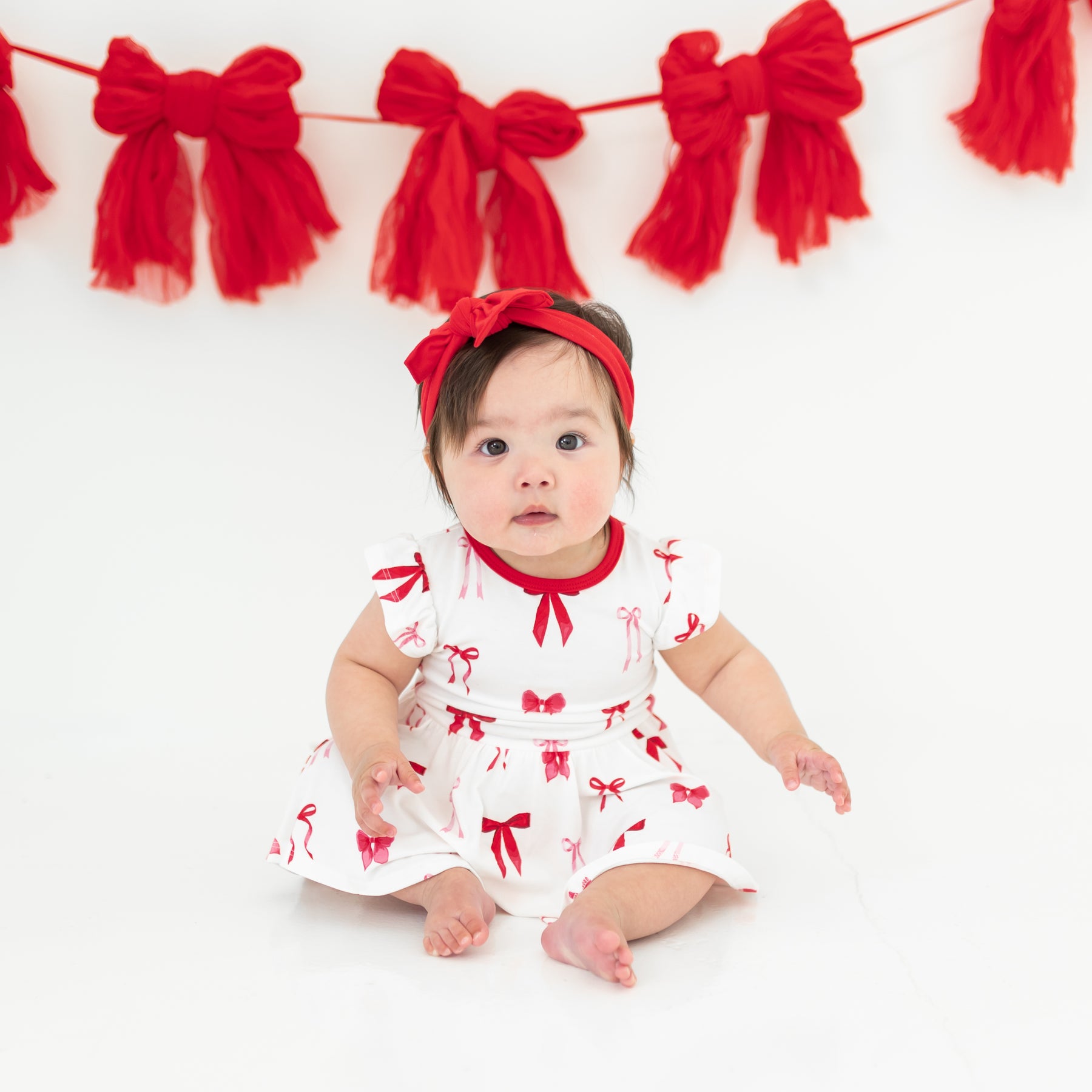 Infant sitting on a light neutral surface wearing the soft and breathable Twirl Bodysuit Dress in Small Love Bow paired with a red bow