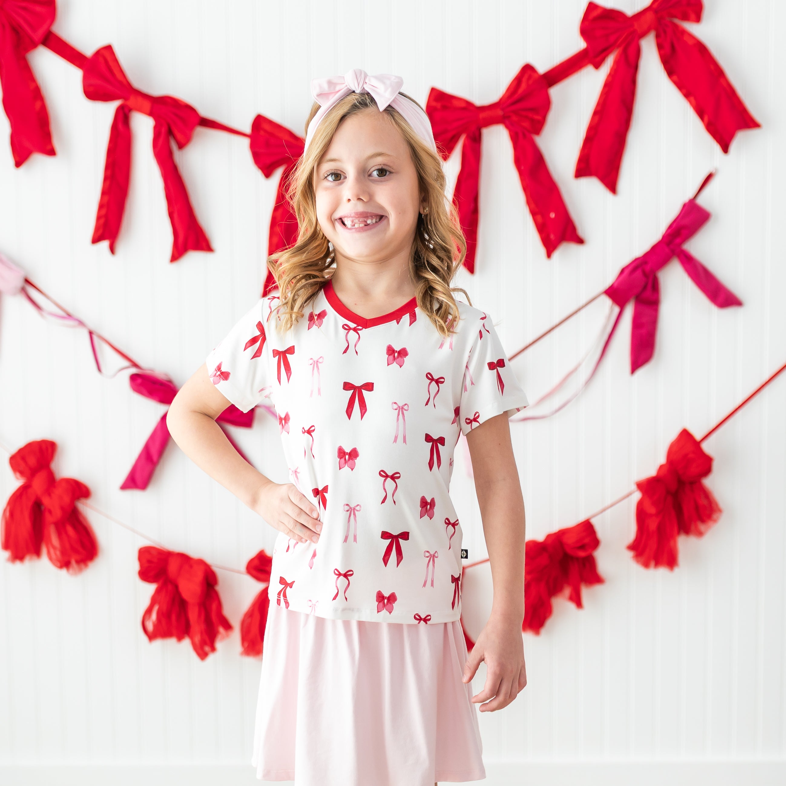 Young girl posing wearing the soft Toddler V-Neck Tee in Small Love Bow paired with a light pink skort