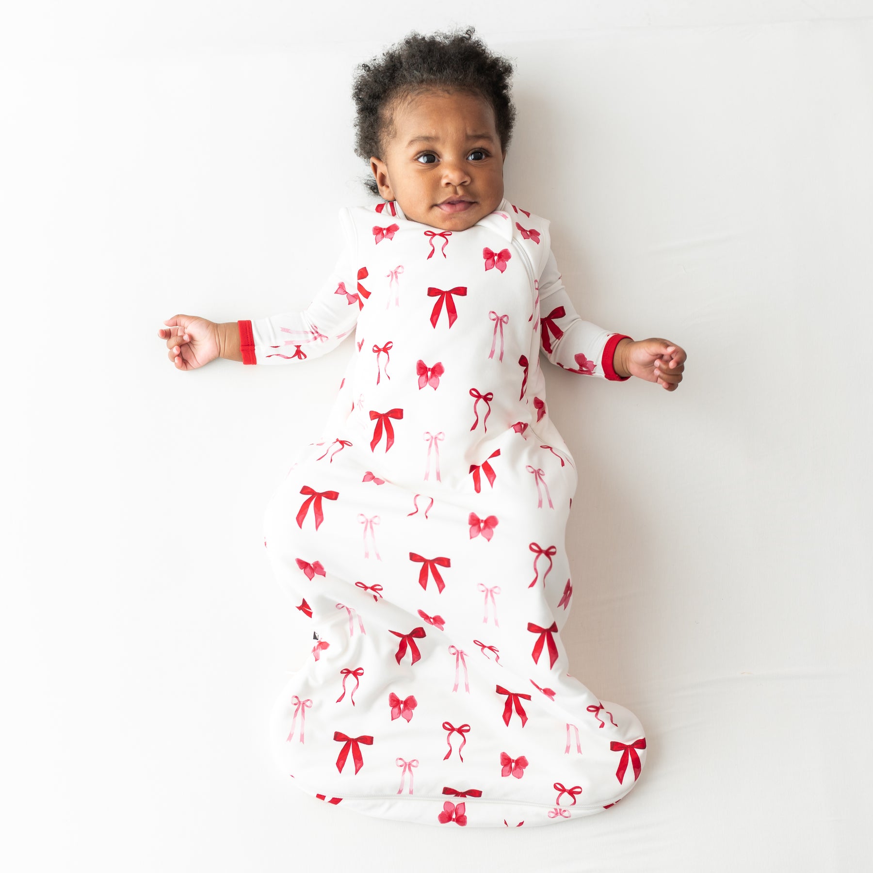 young toddler laying on a light neutral surface wearing the soft and breathable Sleep Bag in Small Love Bow 1.0 with a matching romper on underneath