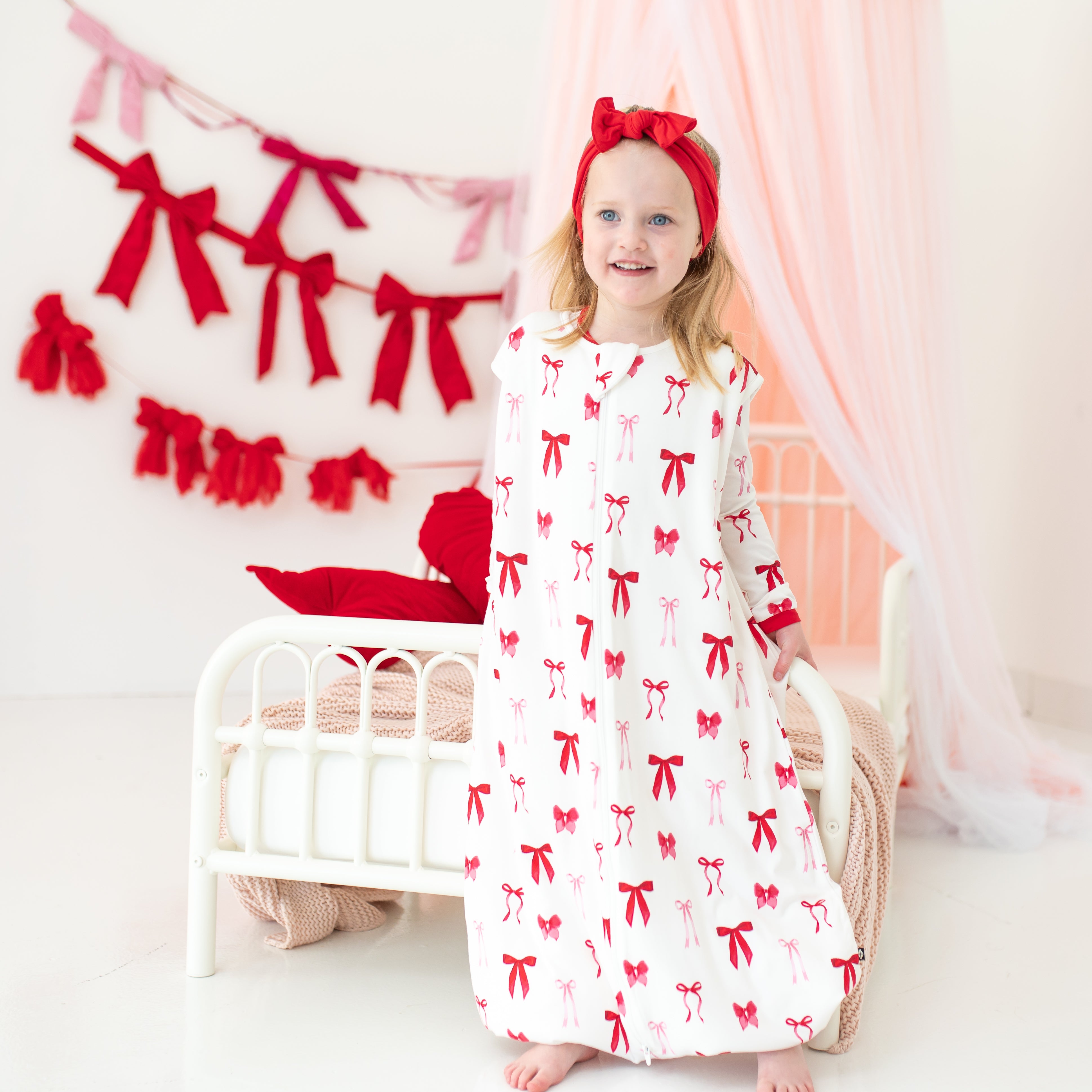 Young girl standing at the end of a bed wearing the soft and breathable Sleep Bag Walker in Small Love Bow 1.0 paired with a Cardinal knotted bow headband