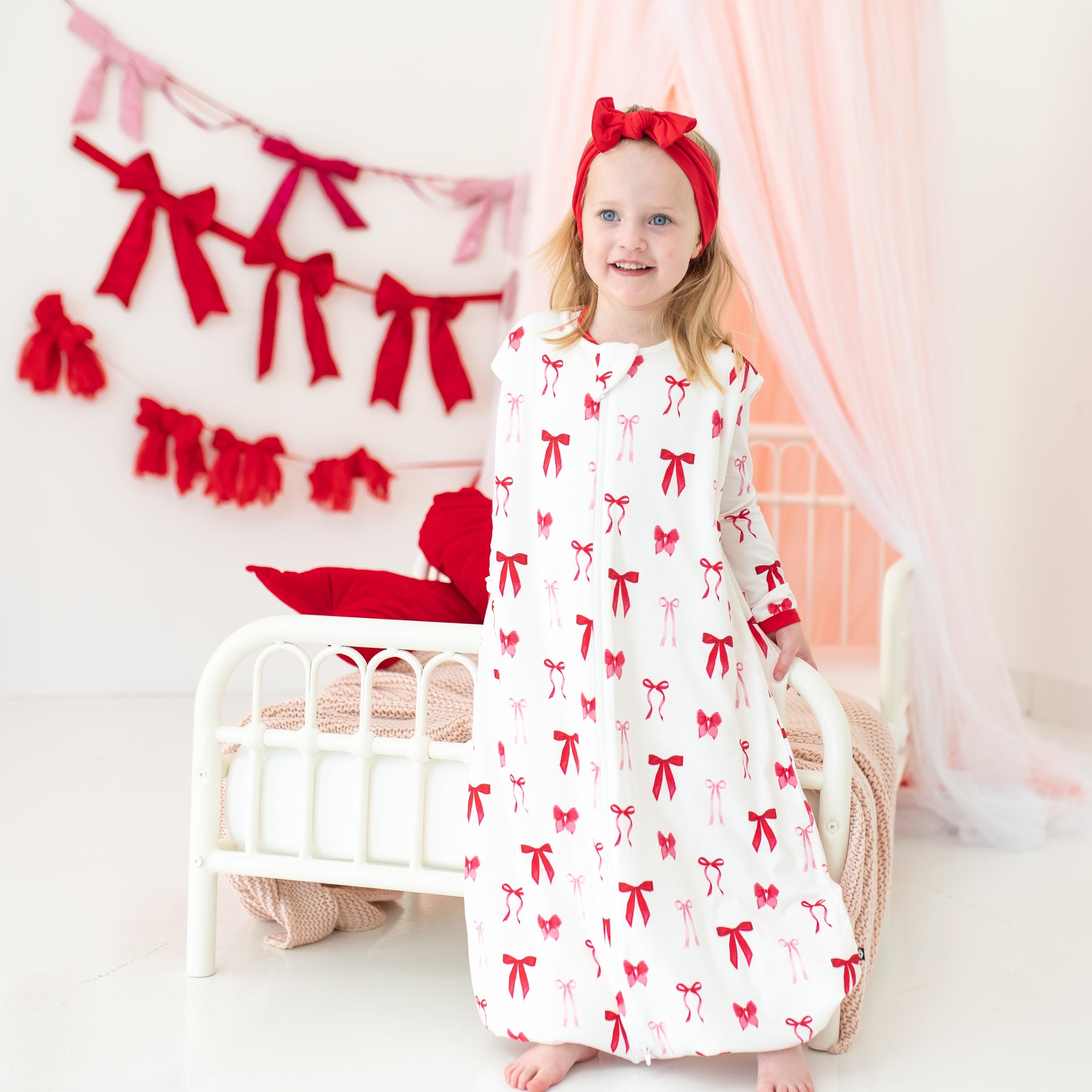 Young girl standing at the end of a bed wearing the soft and breathable Sleep Bag Walker in Small Love Bow 1.0 paired with a Cardinal knotted bow headband