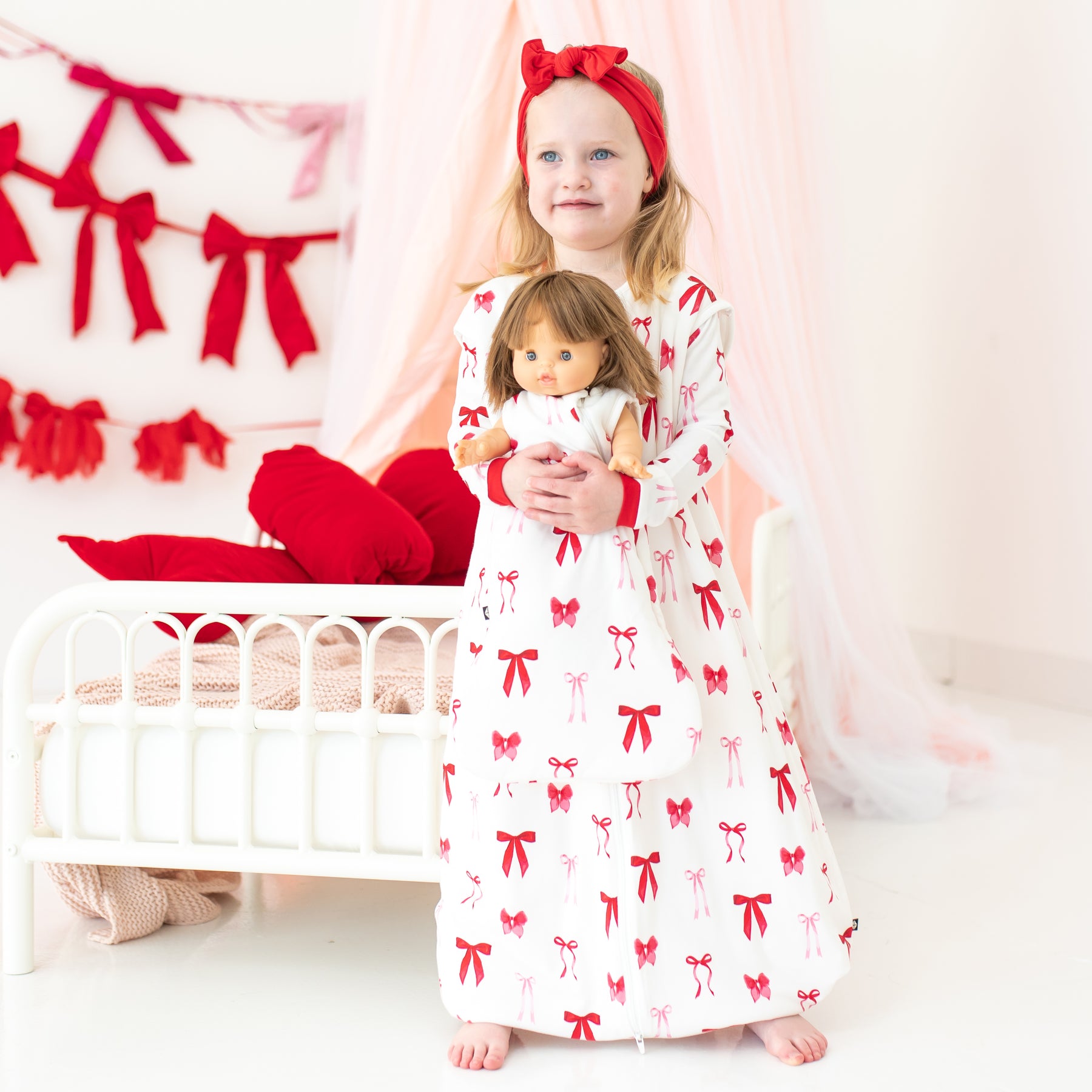 Young girl standing in front of a bed wearing the Sleep Bag Walker in Small Love Bow 1.0 holding a baby doll wearing a matching baby doll sleep bag