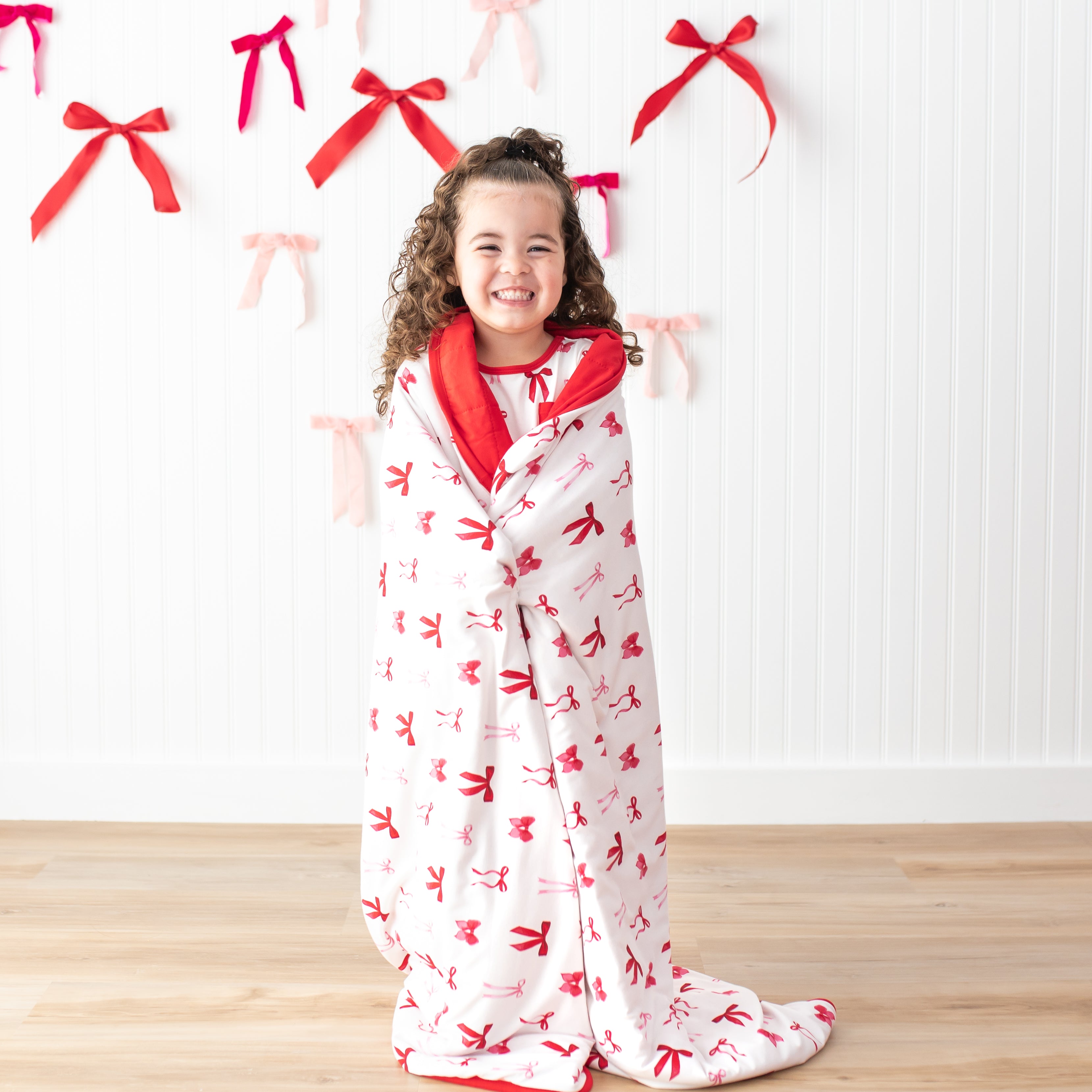 Young smiling girl standing in front of a white wall wearing the Toddler Blanket in Small Love Bow 1.0 around her shoulders