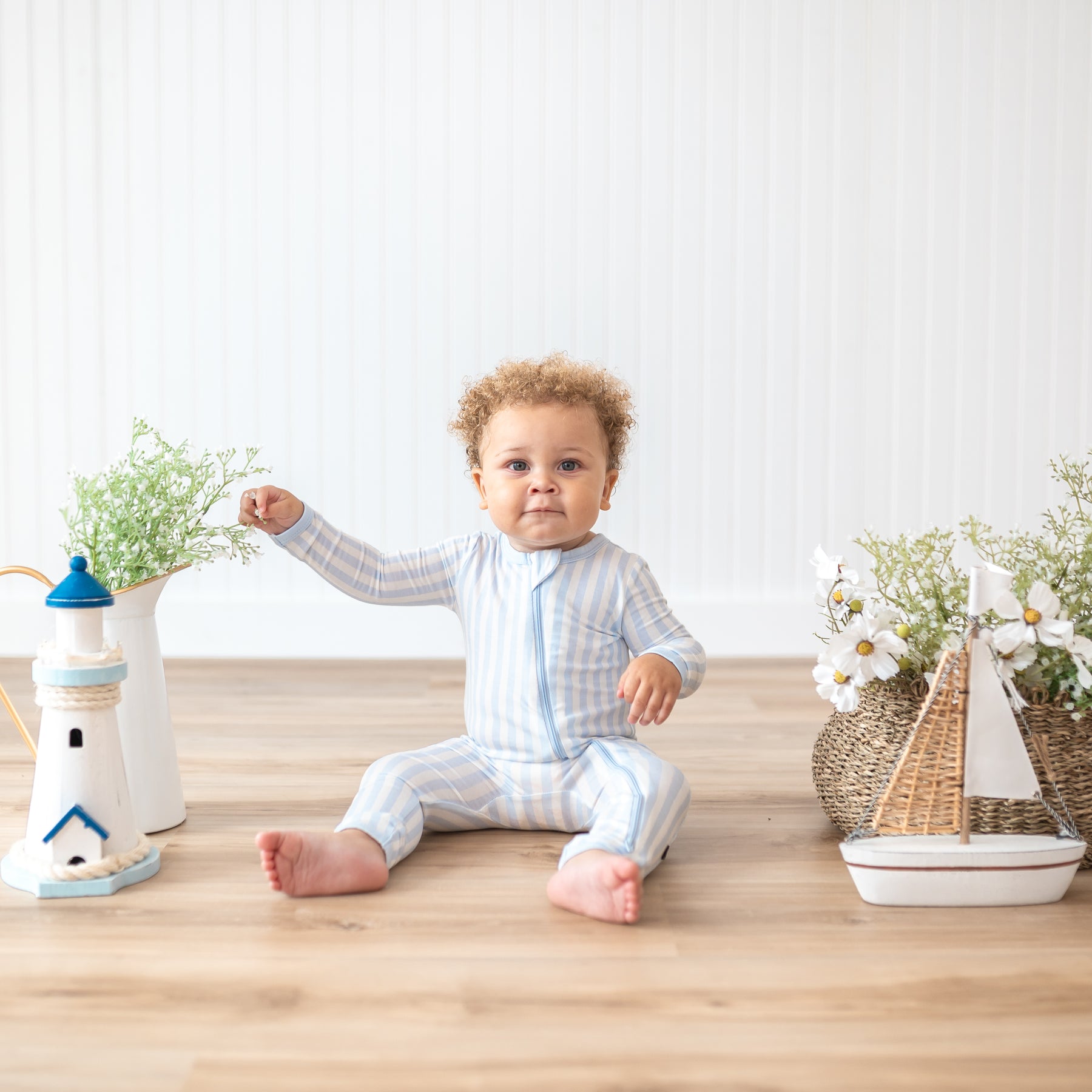 Toddler sitting down touching the potted plants wearing the Zippered Romper in Small Mist Stripe