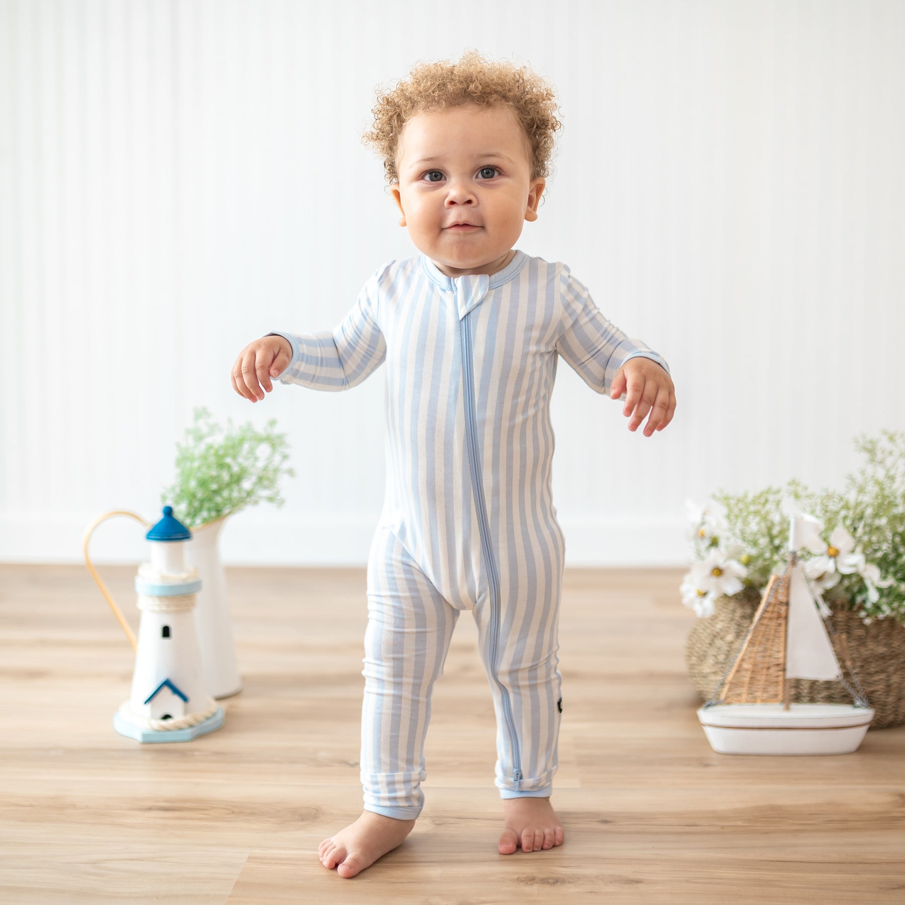 Toddler standing in front of nautical themed props and potted plants and flowers wearing the Zippered Romper in Small Mist Stripe