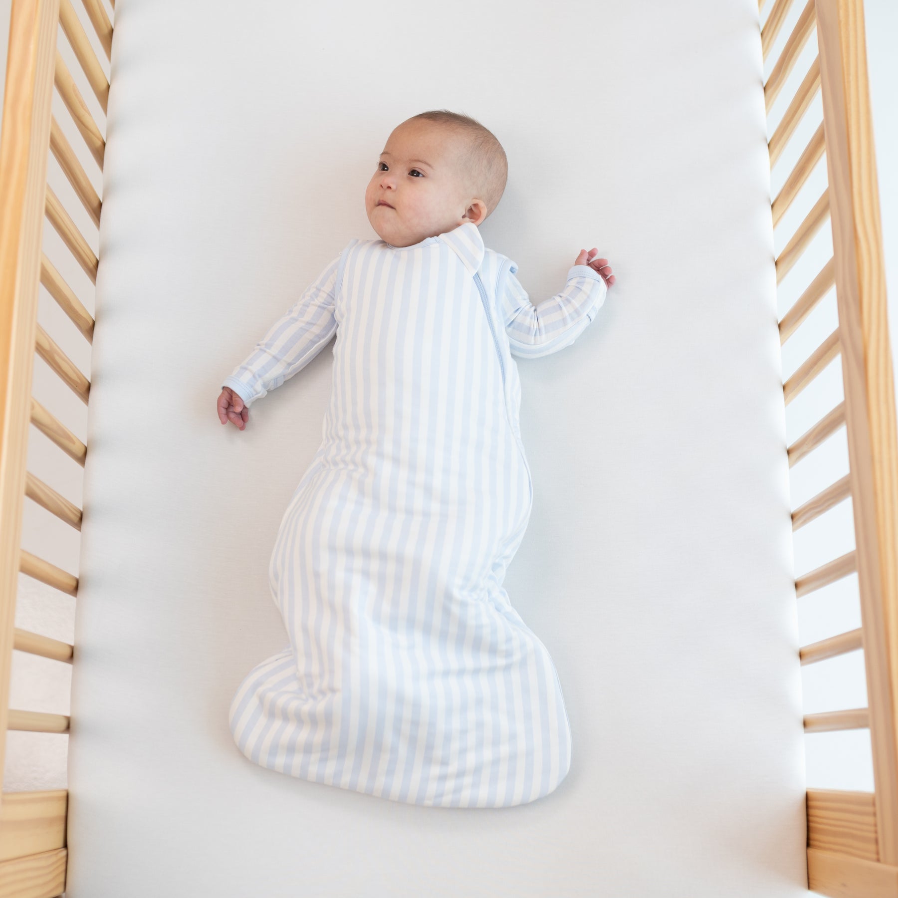 Infant laying in a crib on a Cloud Crib Sheet wearing the Sleep Bag in Small Mist Stripe 1.0 with matching long sleeve romper