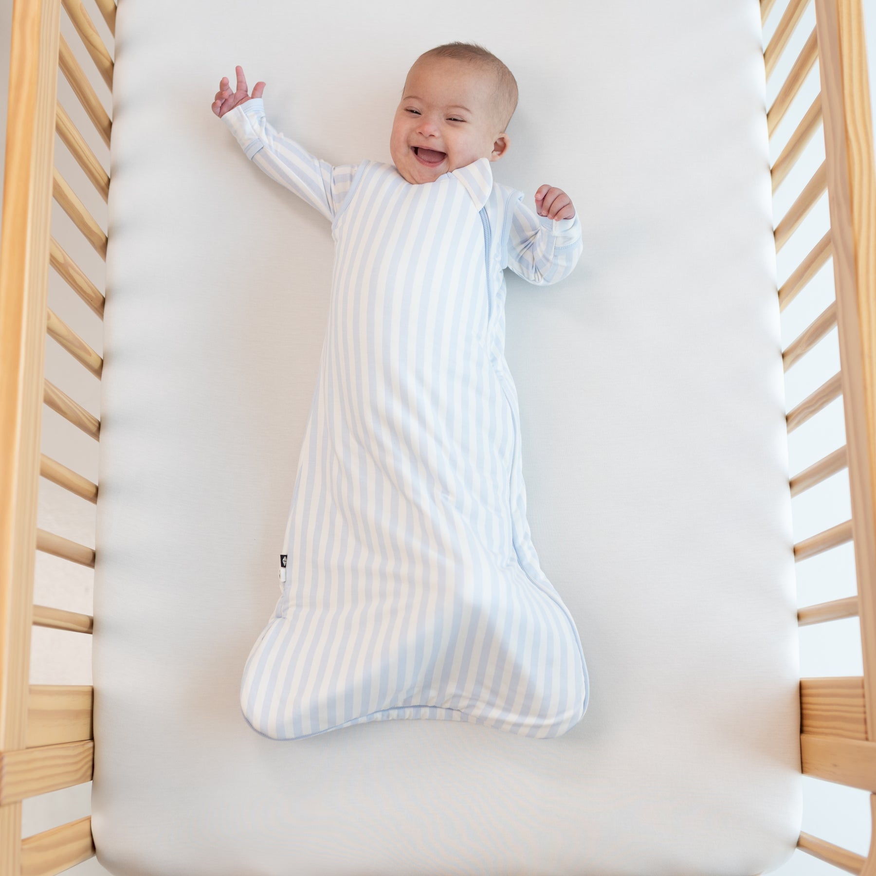 Smiling infant laying in a crib with one hand above their head wearing the Sleep Bag in Small Mist Stripe 1.0 with matching long sleeve romper