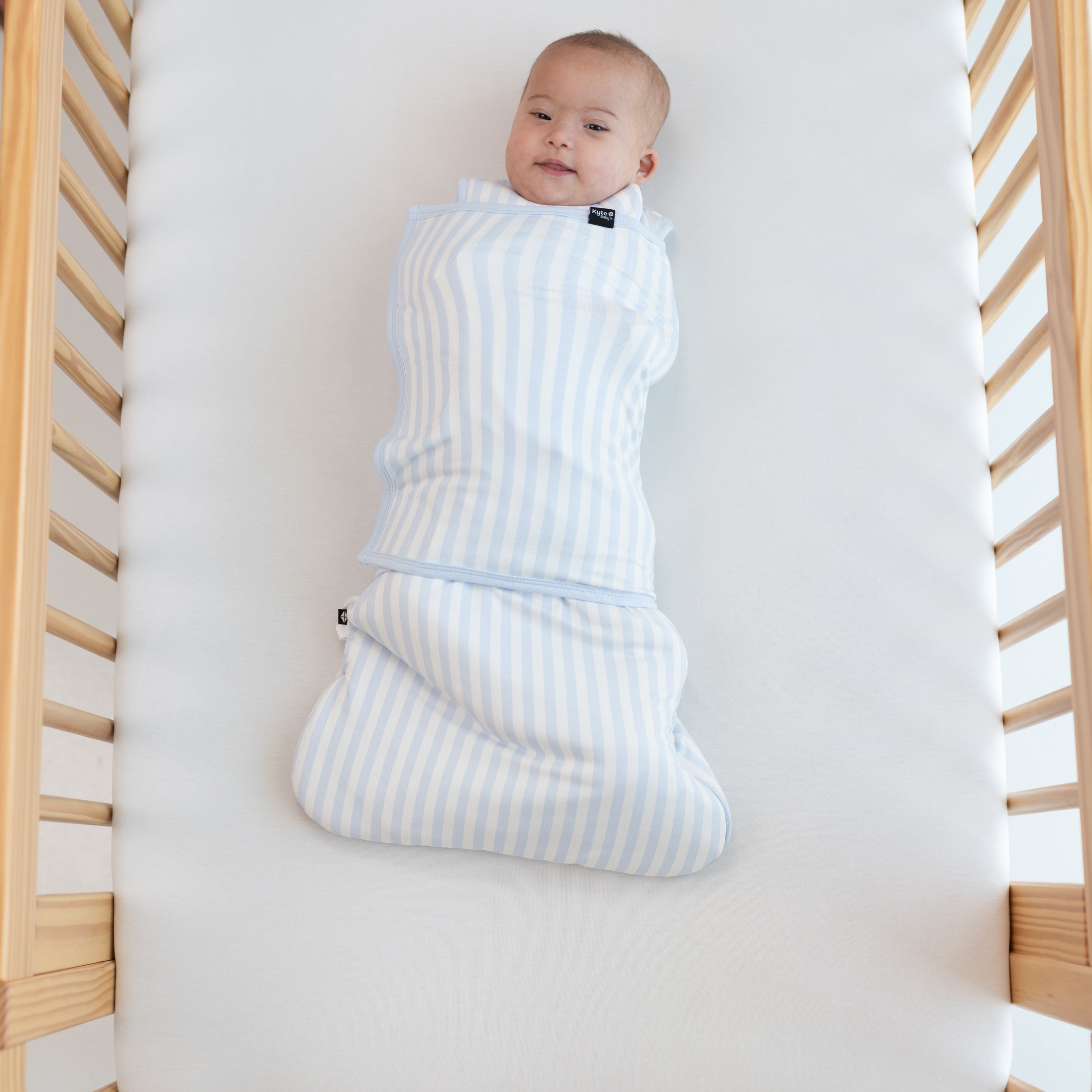 Smiling infant laying in a crib swaddled in the Sleep Bag Swaddler in Small Mist Stripe 1.0