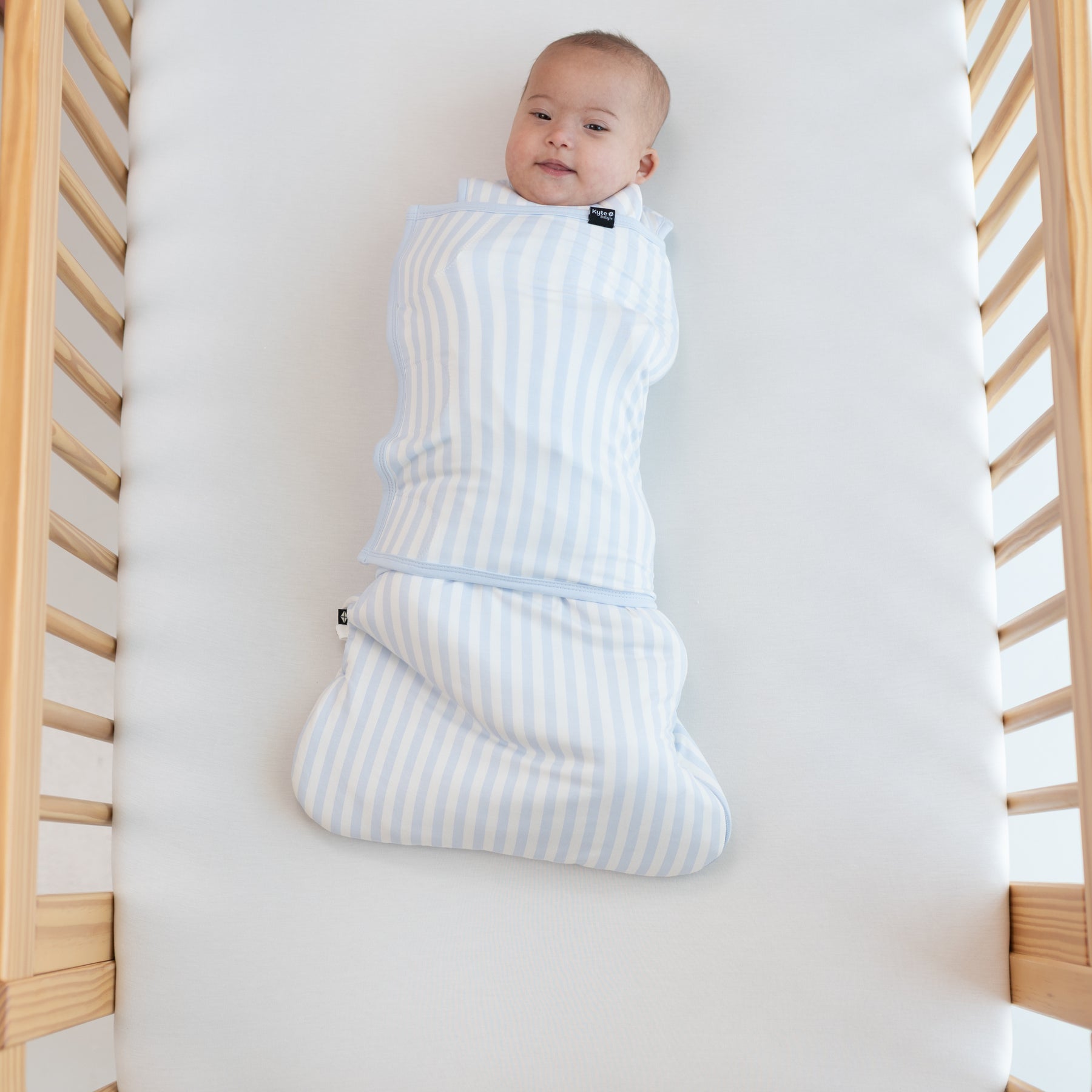 Smiling infant laying in a crib swaddled in the Sleep Bag Swaddler in Small Mist Stripe 1.0