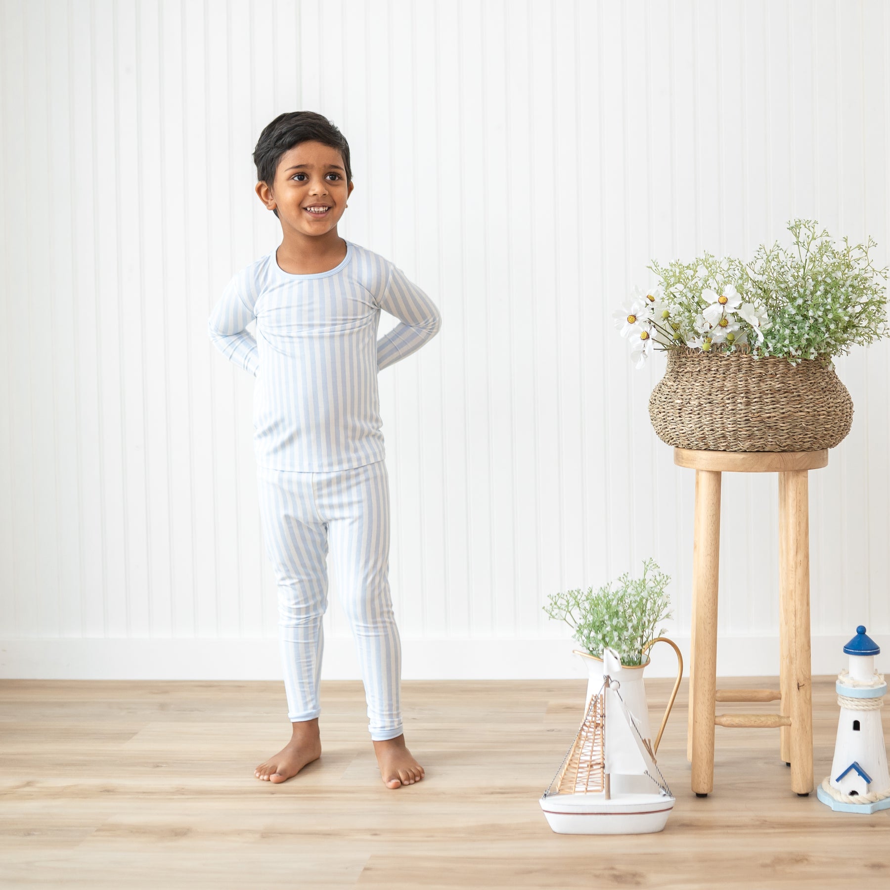 Boy modeling the Long Sleeve Pajamas in Small Mist Stripe beside potted flowers and a nautical themed toys
