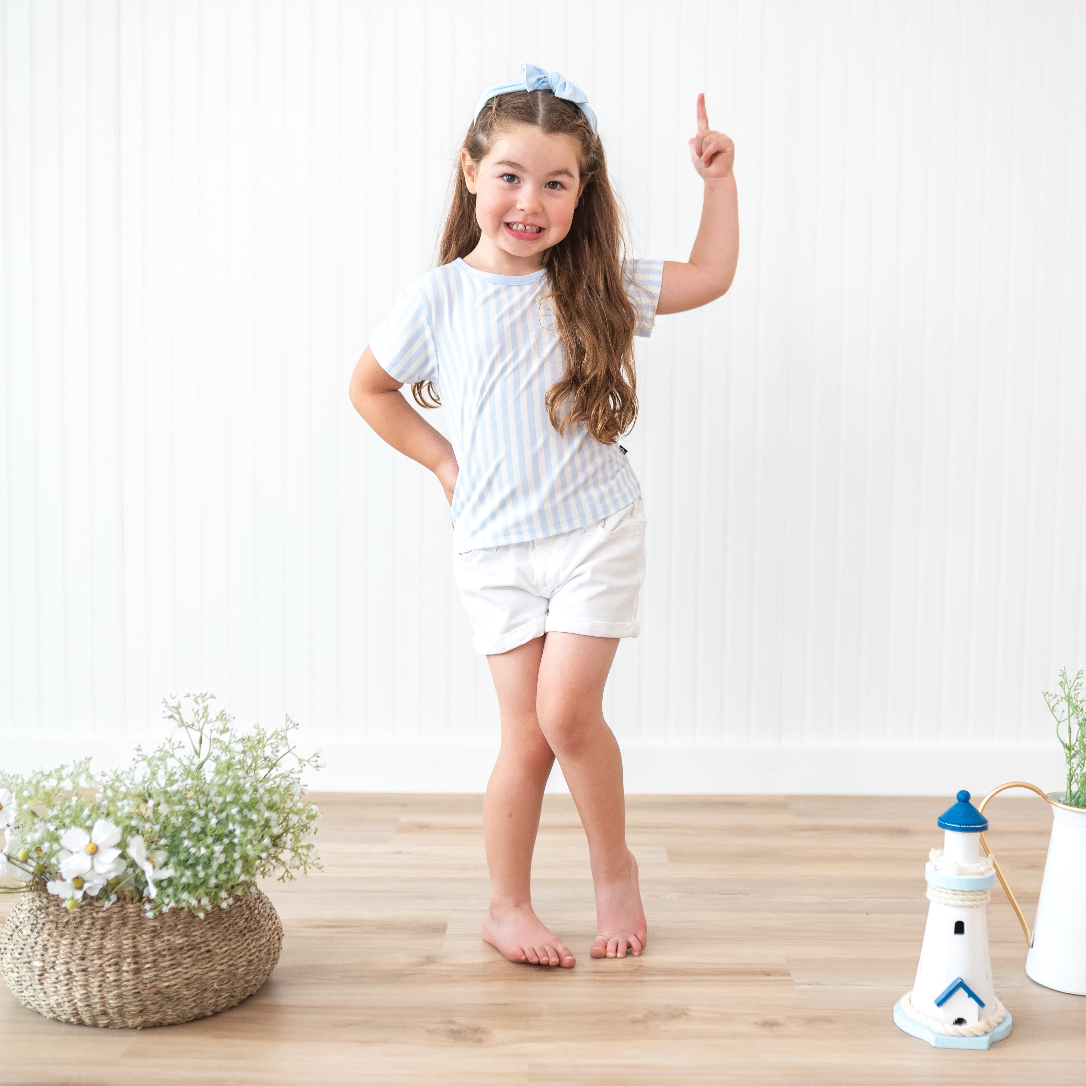 Toddler girl standing with one hand on her hip and the other hand pointing upwards wearing the Toddler Basic Tee in Small Mist Stripe paired with white shorts