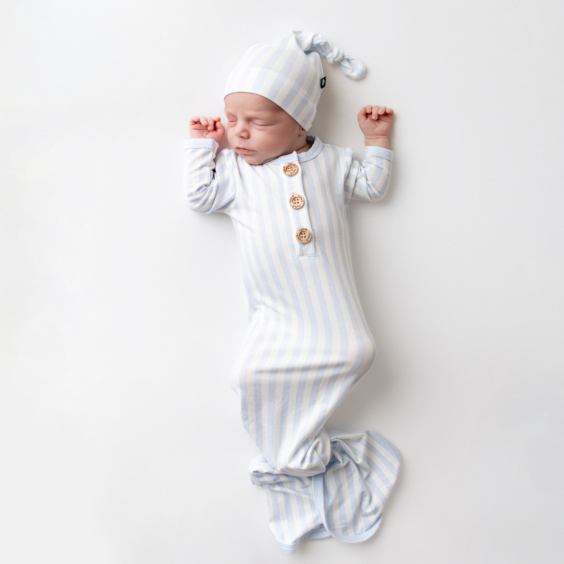 Sleeping newborn laying on a light neutral surface wearing the Knotted Gown with Hat Set in Small Mist Stripe