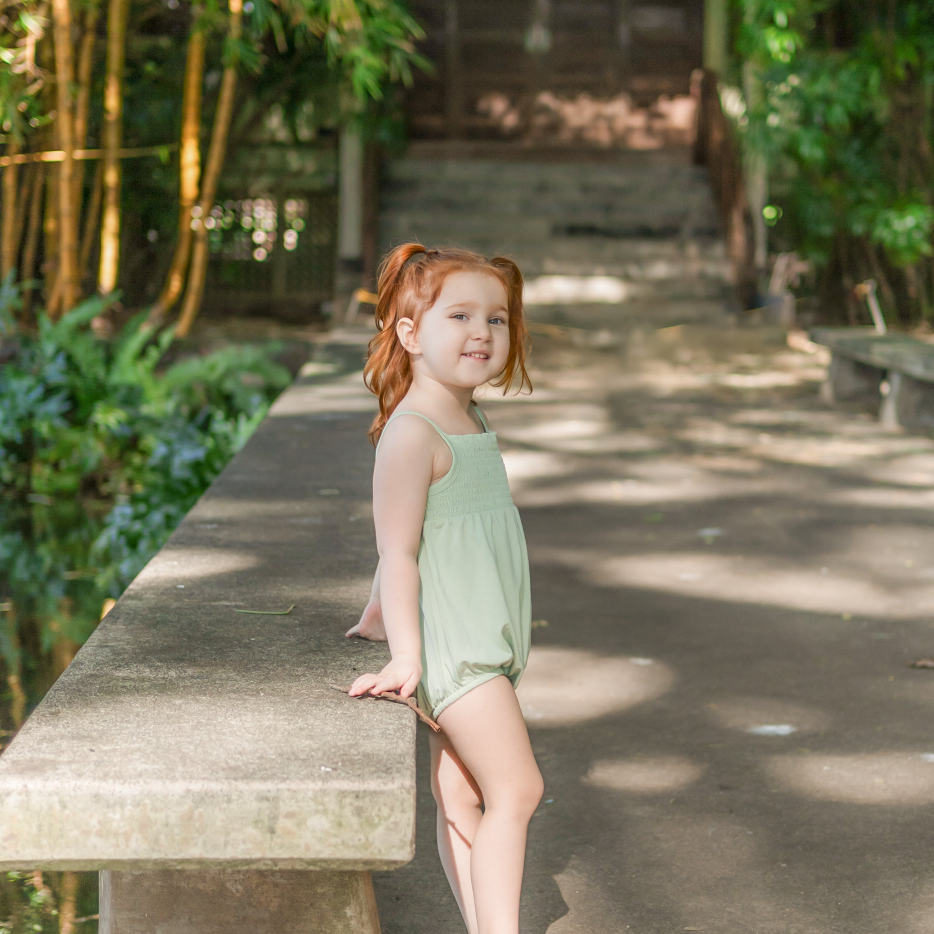 Young girl leaning against a concreted bench wearing the Smocked Bubble Romper in Basil