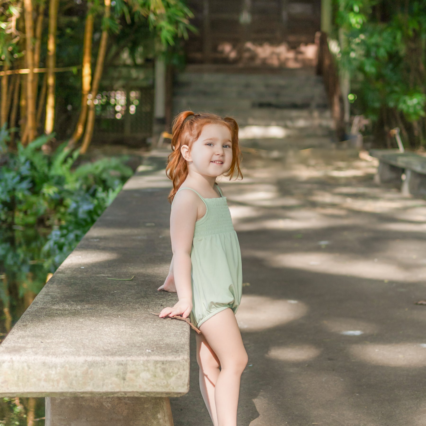 Young girl leaning against a concreted bench wearing the Smocked Bubble Romper in Basil