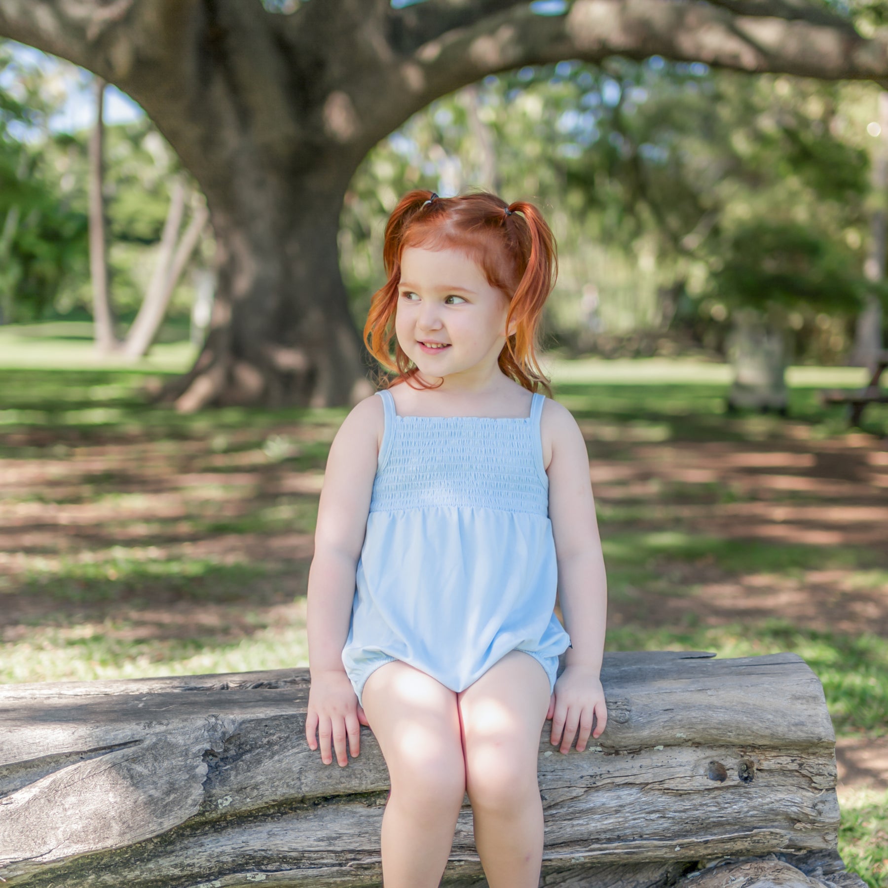 Young girl sitting in a park on a wooden log wearing the Smocked Bubble Romper in Breeze