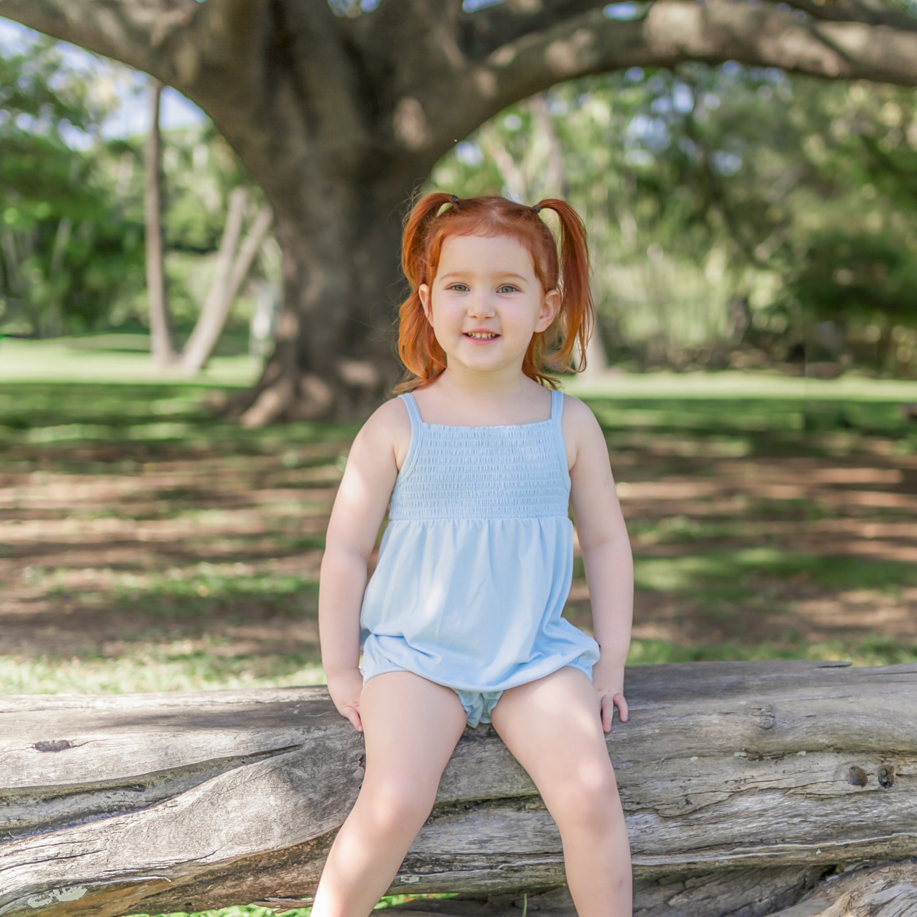 Smiling girl sitting on a wooden log in a park wearing the Smocked Bubble Romper in Breeze