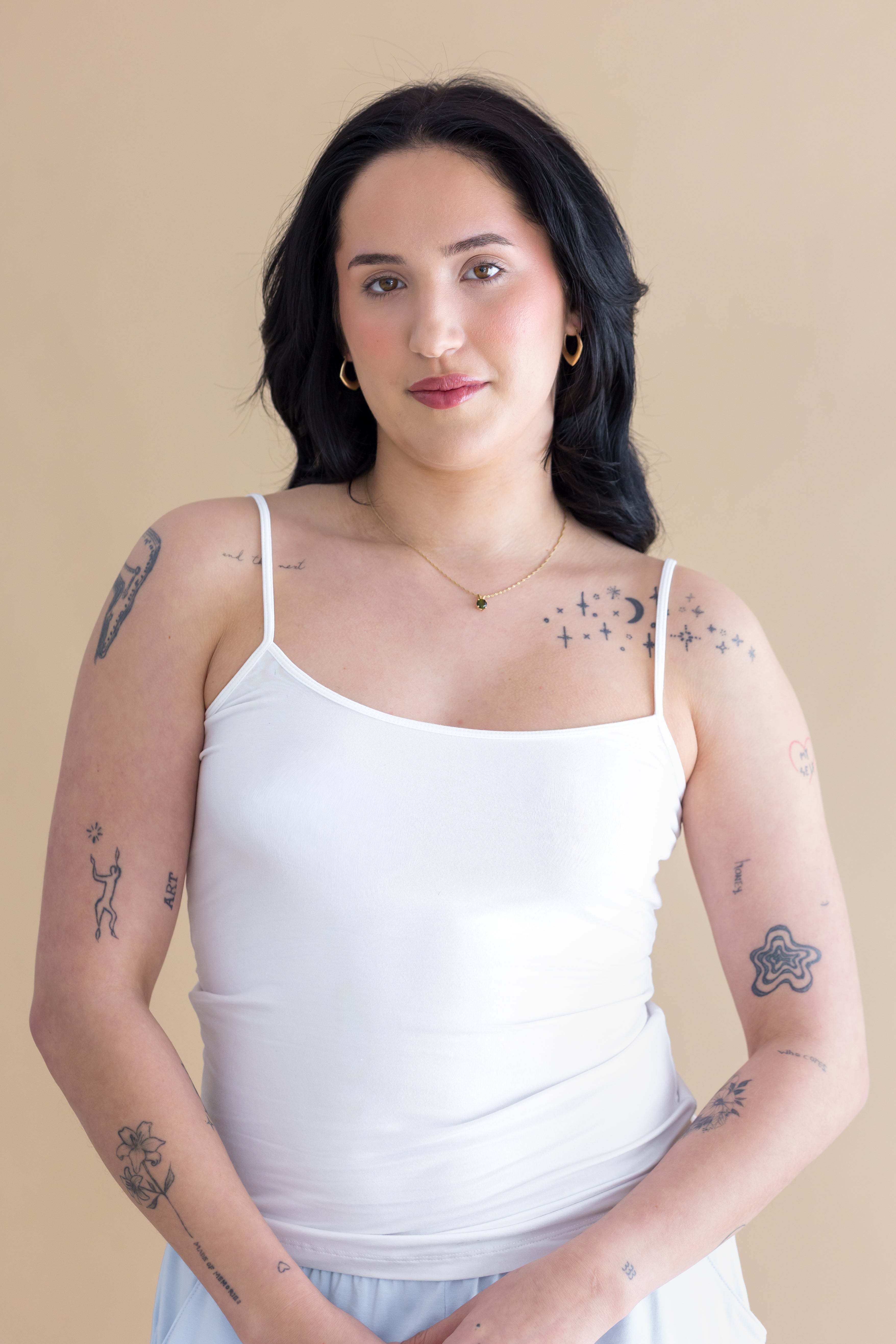 Woman wearing a white tank top with tattoos on her arms against a beige background