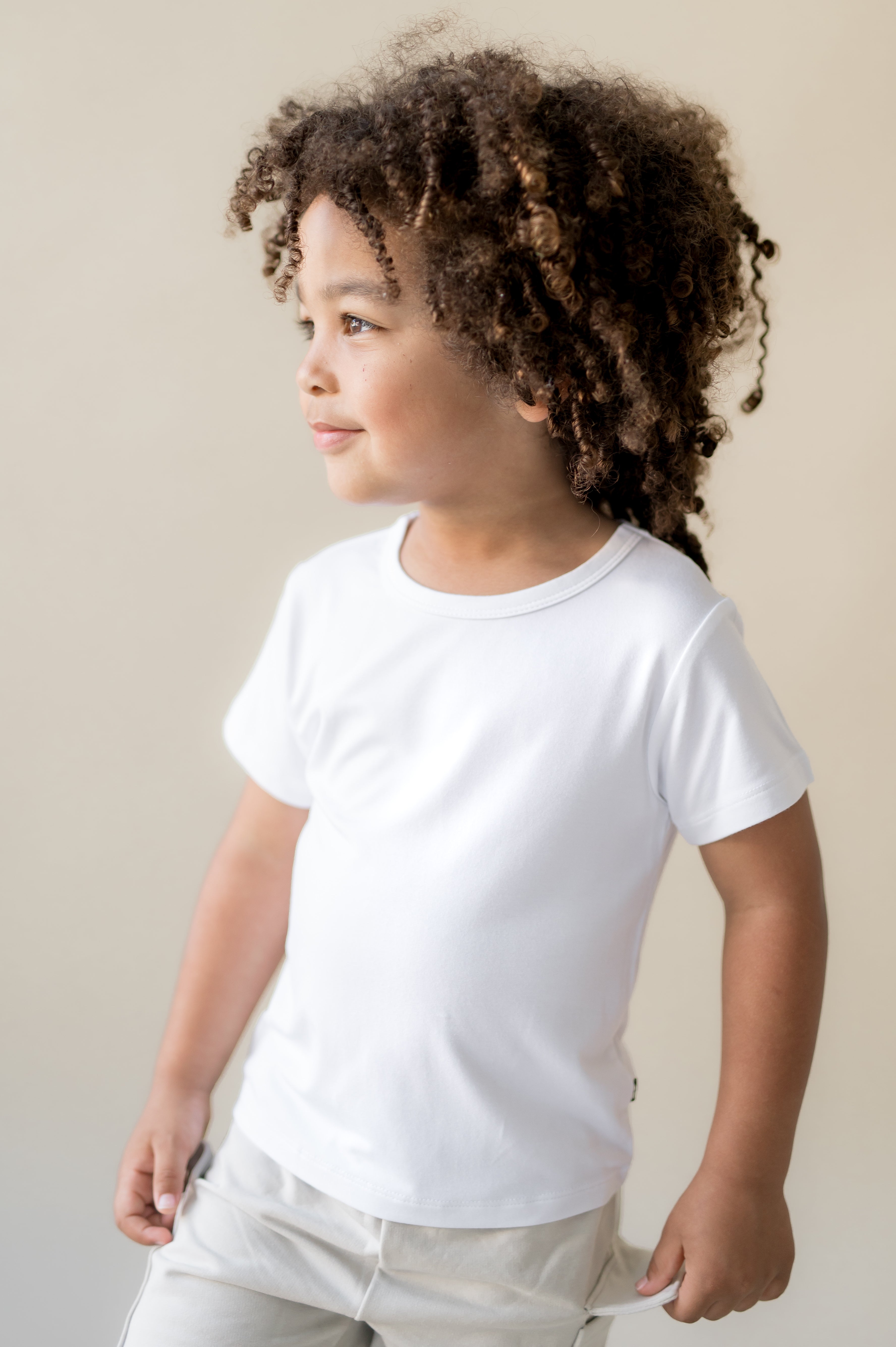 Young boy looking off to the side wearing the Toddler Basic Tee in Snow with light colored pants