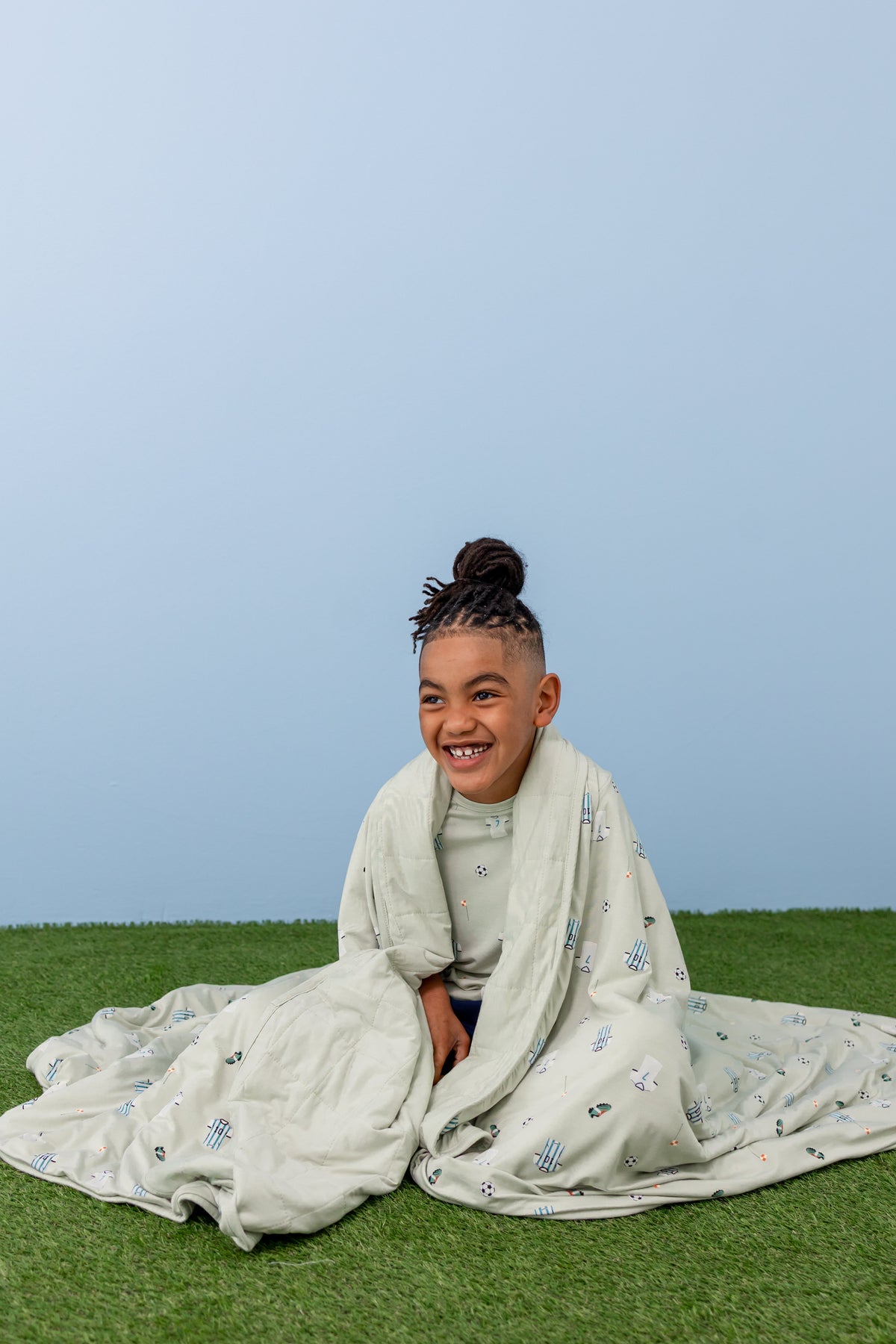 Child wrapped in Soccer print Youth blanket wearing a similar print Tee sitting on a green grass