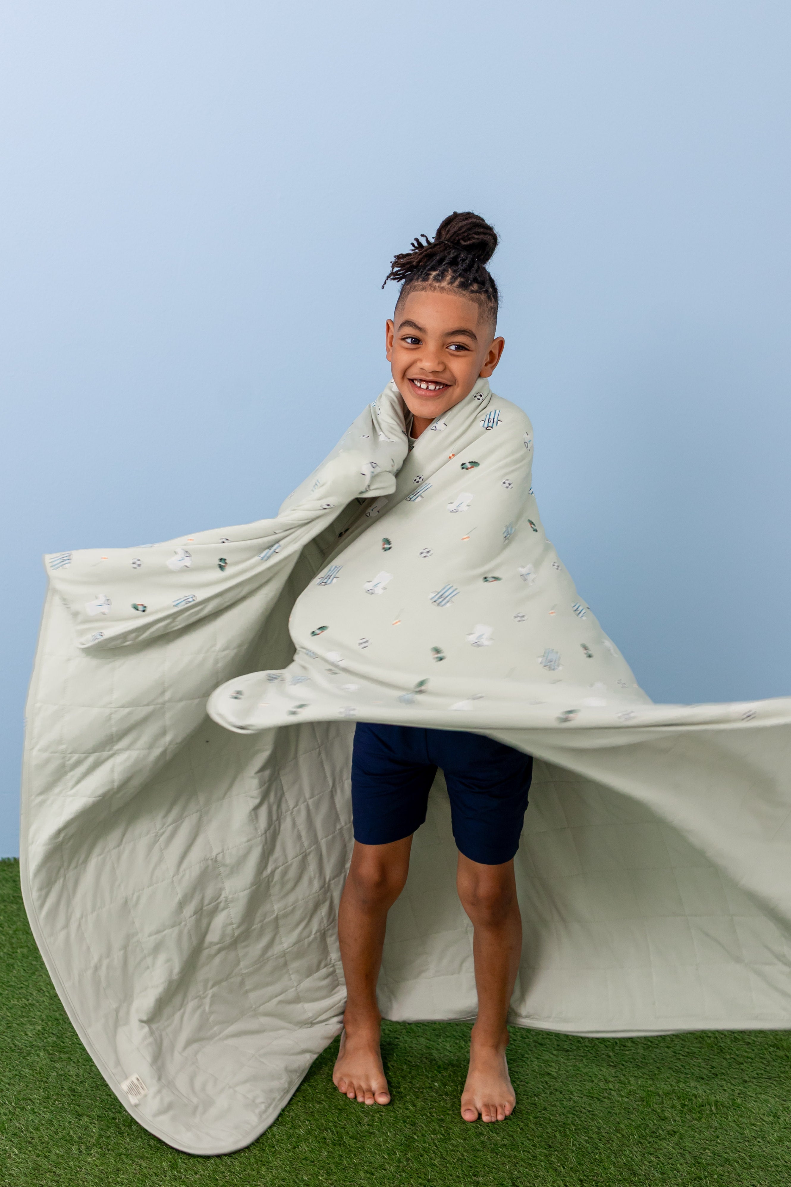 Child standing wrapped in Soccer print Youth blanket wearing navy shorts on a green grass