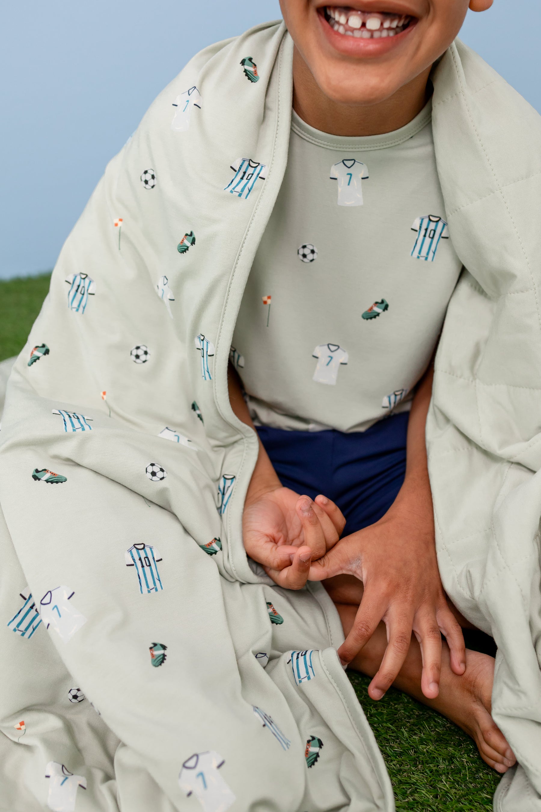 Child smiling with Soccer print Youth blanket wearing a similar print Tee and navy shorts sitting on a green grass