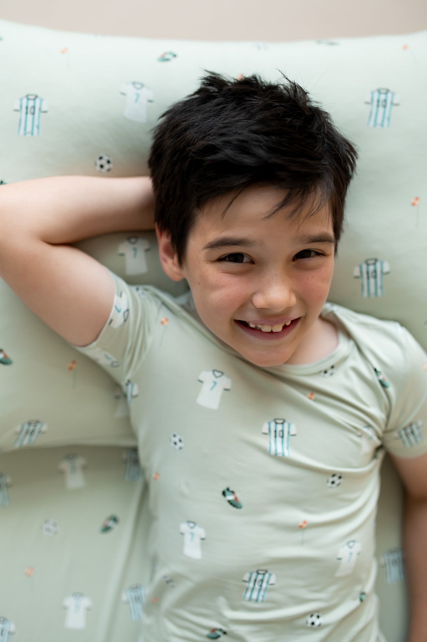 Child wearing a light-colored shirt with small soccer patterns, lying on a cushion with a patterned design.