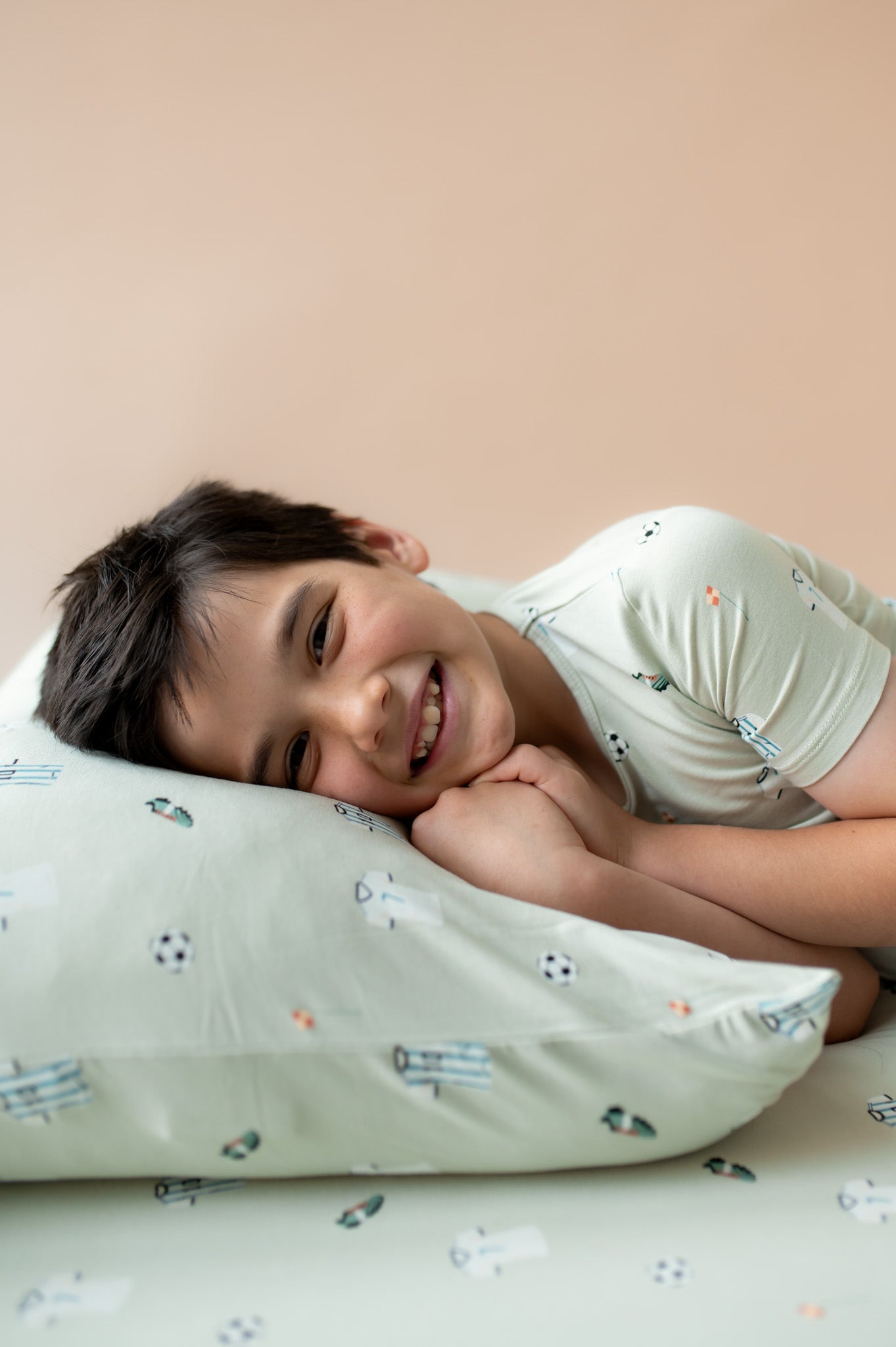 Child lying on a pillow with a pattern of small soccer balls and jerseys against a beige wall.