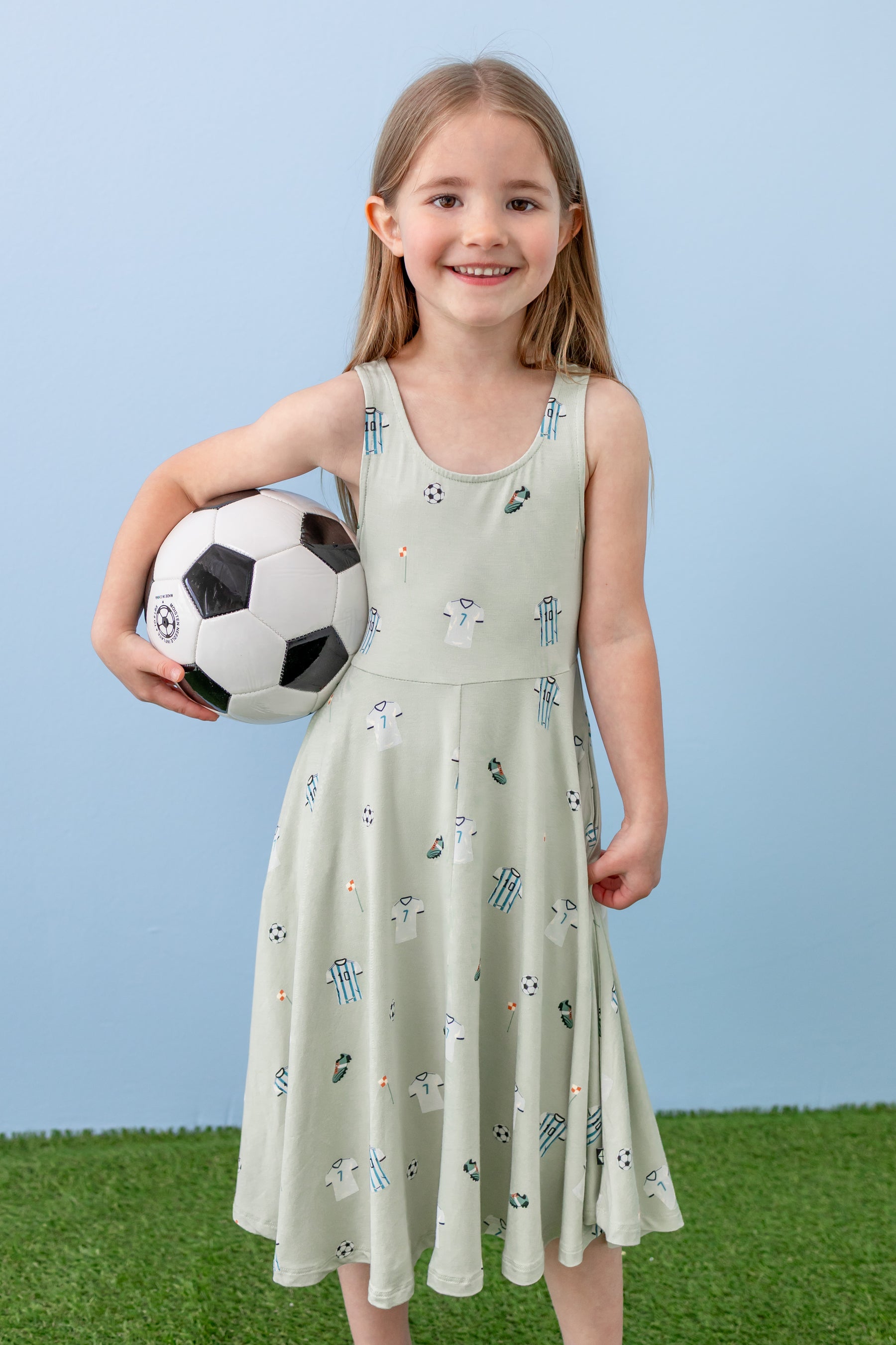 Child wearing Tank Twirl dress in Soccer print holding a soccer ball on green grass