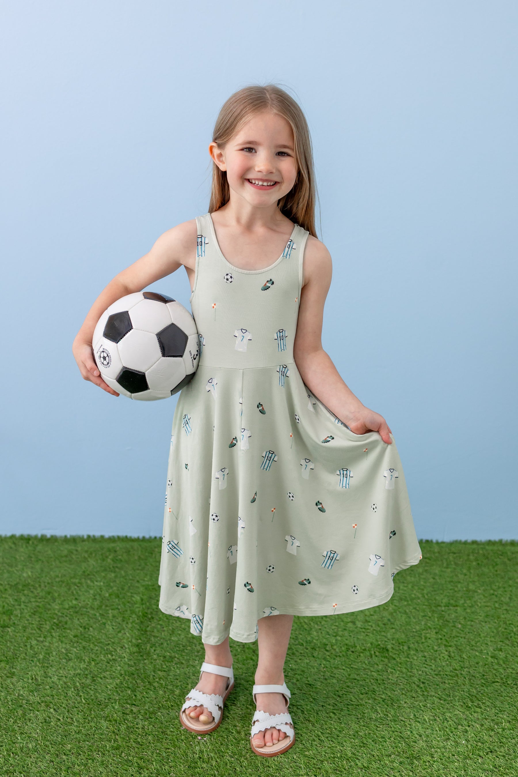 Child posing in Tank Twirl dress in Soccer print holding a soccer ball on green grass