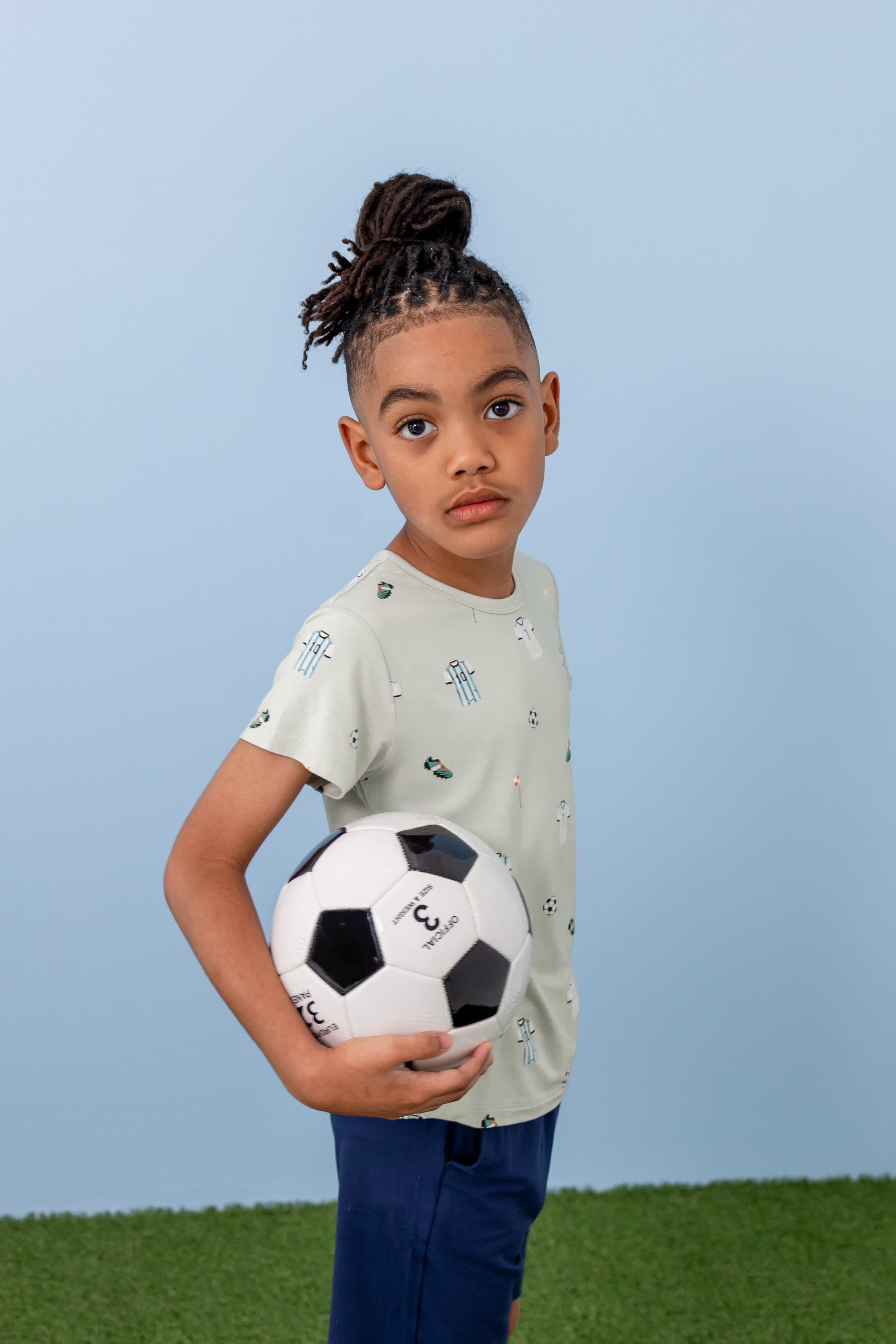 Child wearing Toddler Basic Tee in Soccer print over navy shorts on green grass holding a soccer ball