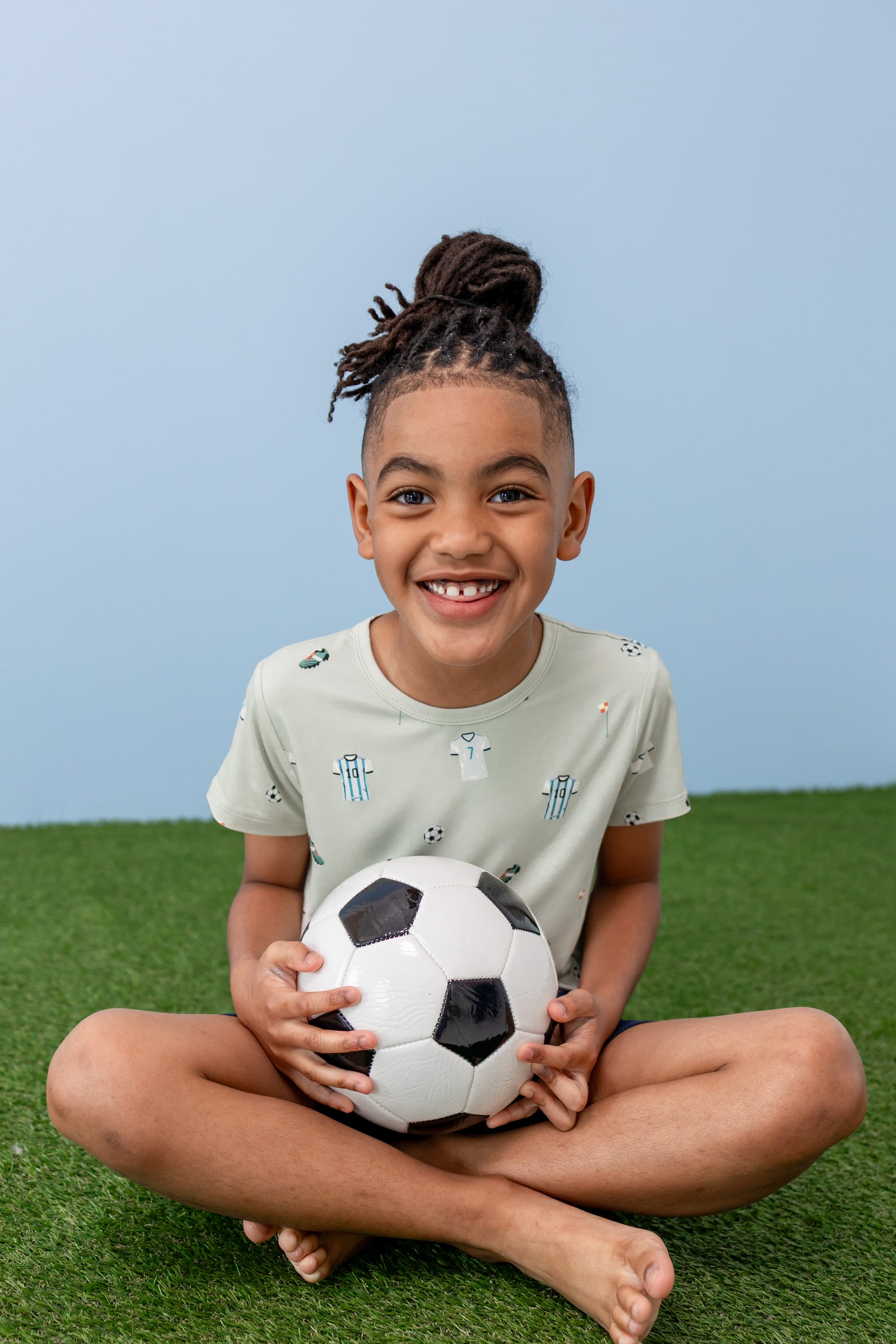Child sitting in Toddler Basic Tee in Soccer print on green grass with a soccer ball