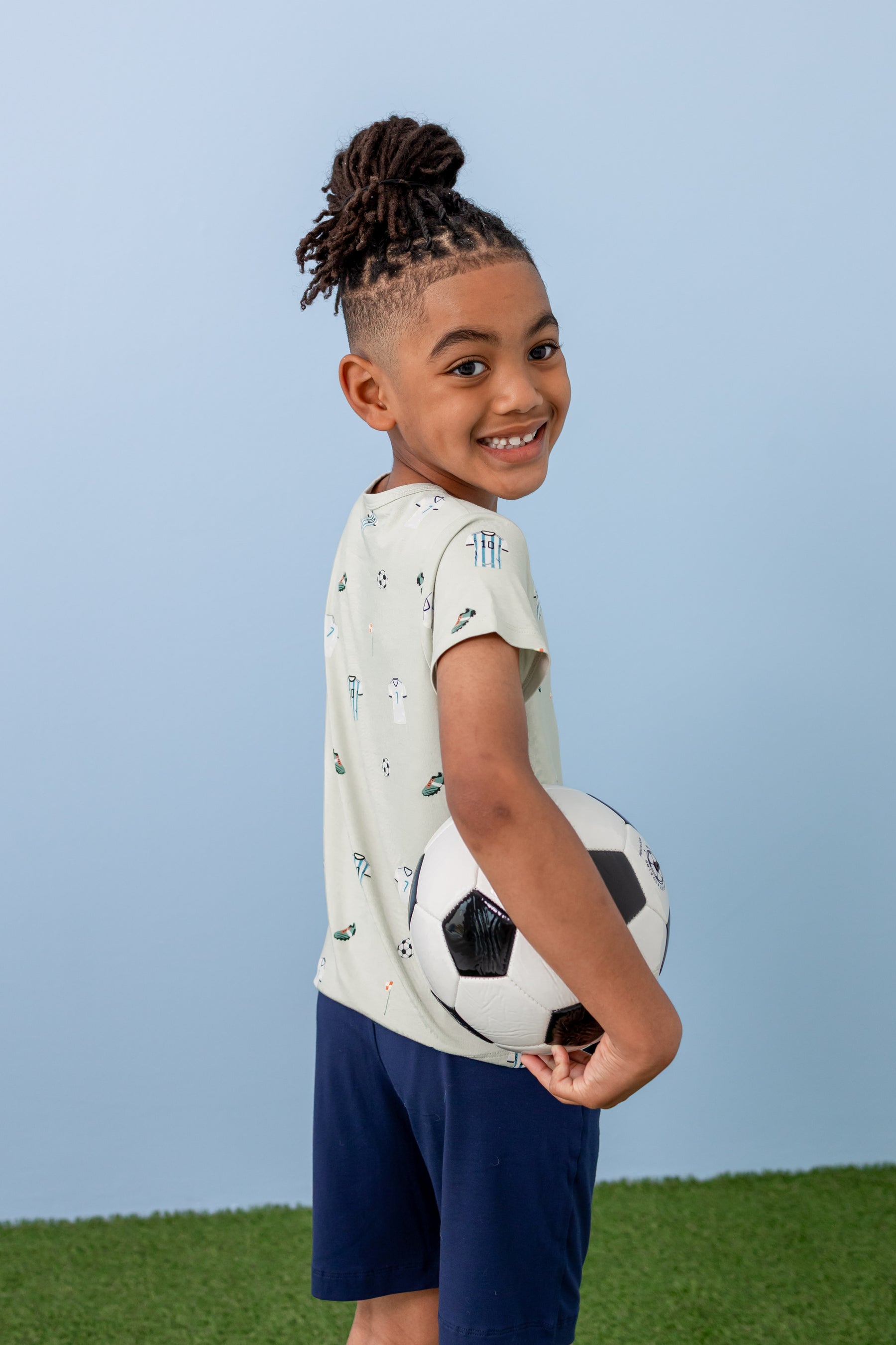 Child posing in Toddler Basic Tee in Soccer print over navy shorts on green grass holding a soccer ball