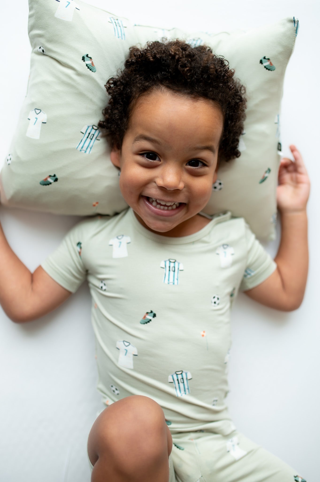 Child wearing a light green soccer pajamas, lying on a white surface with a soccer patterned pillow.