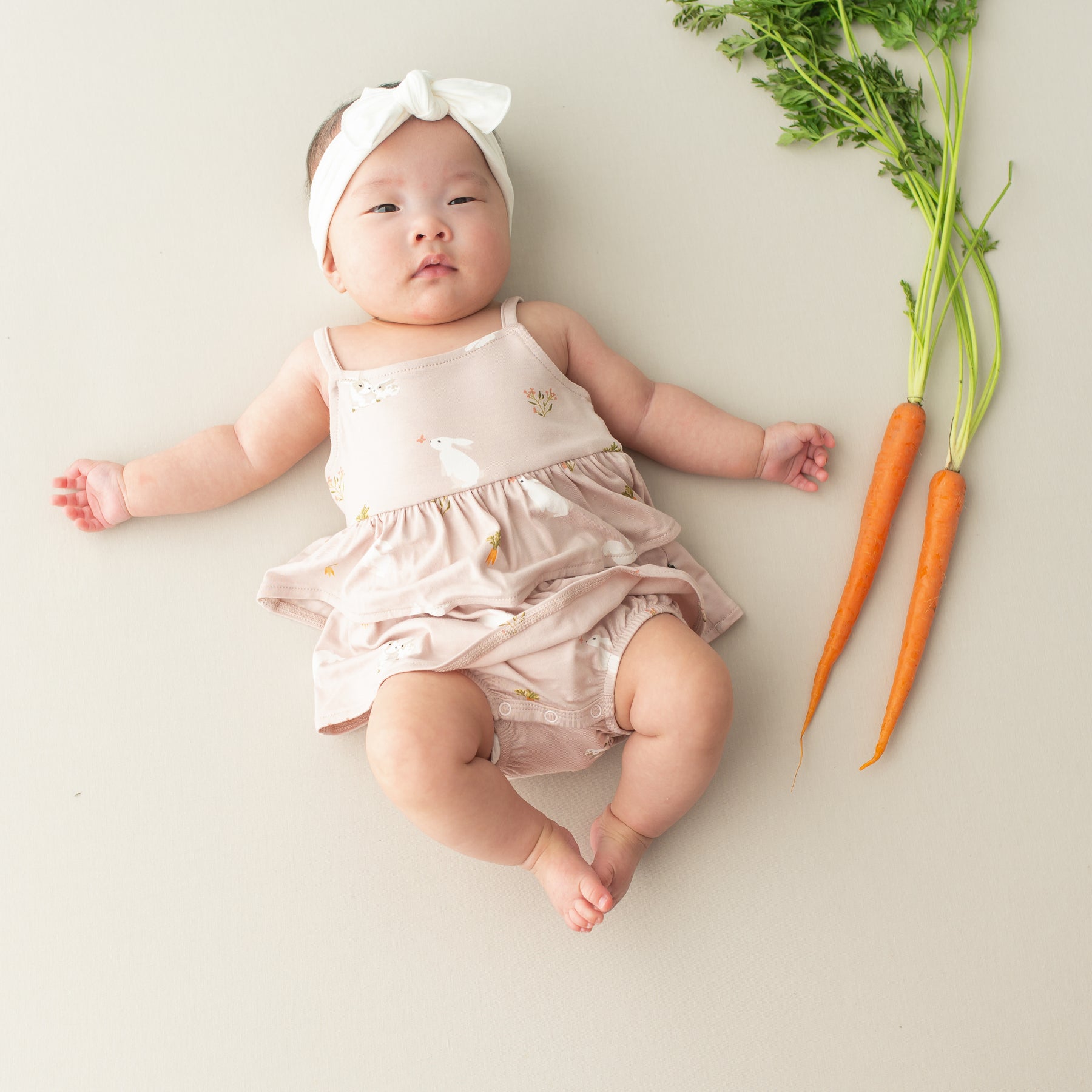 Baby wearing Spaghetti Strap Ruffle Romper in Blush Rabbit next to carrots