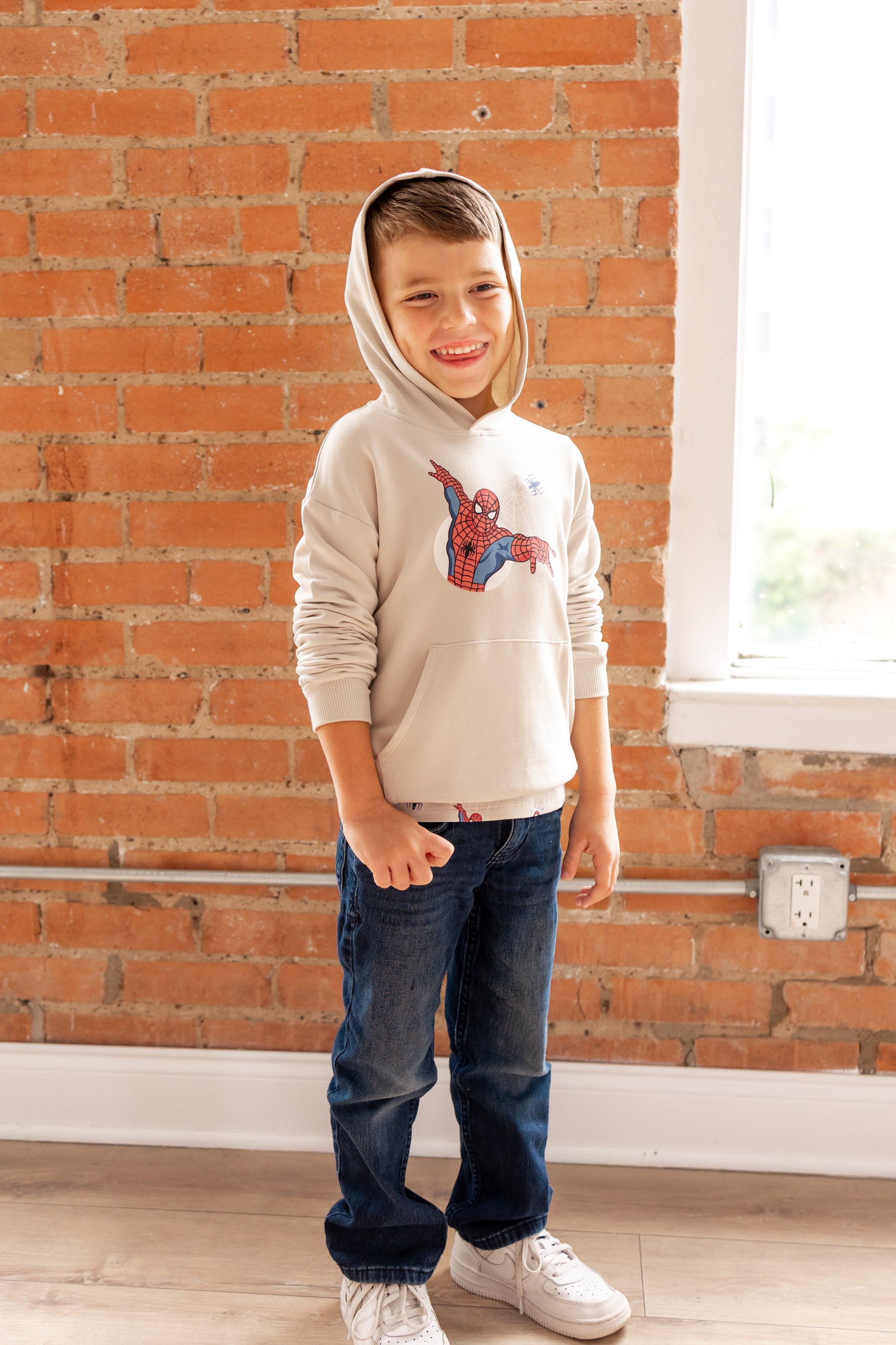 Boy smiling with an oat color hoodie cap on his head in front of a brick wall with a Spider-Man print in center over a blue jeans and white sneakers
