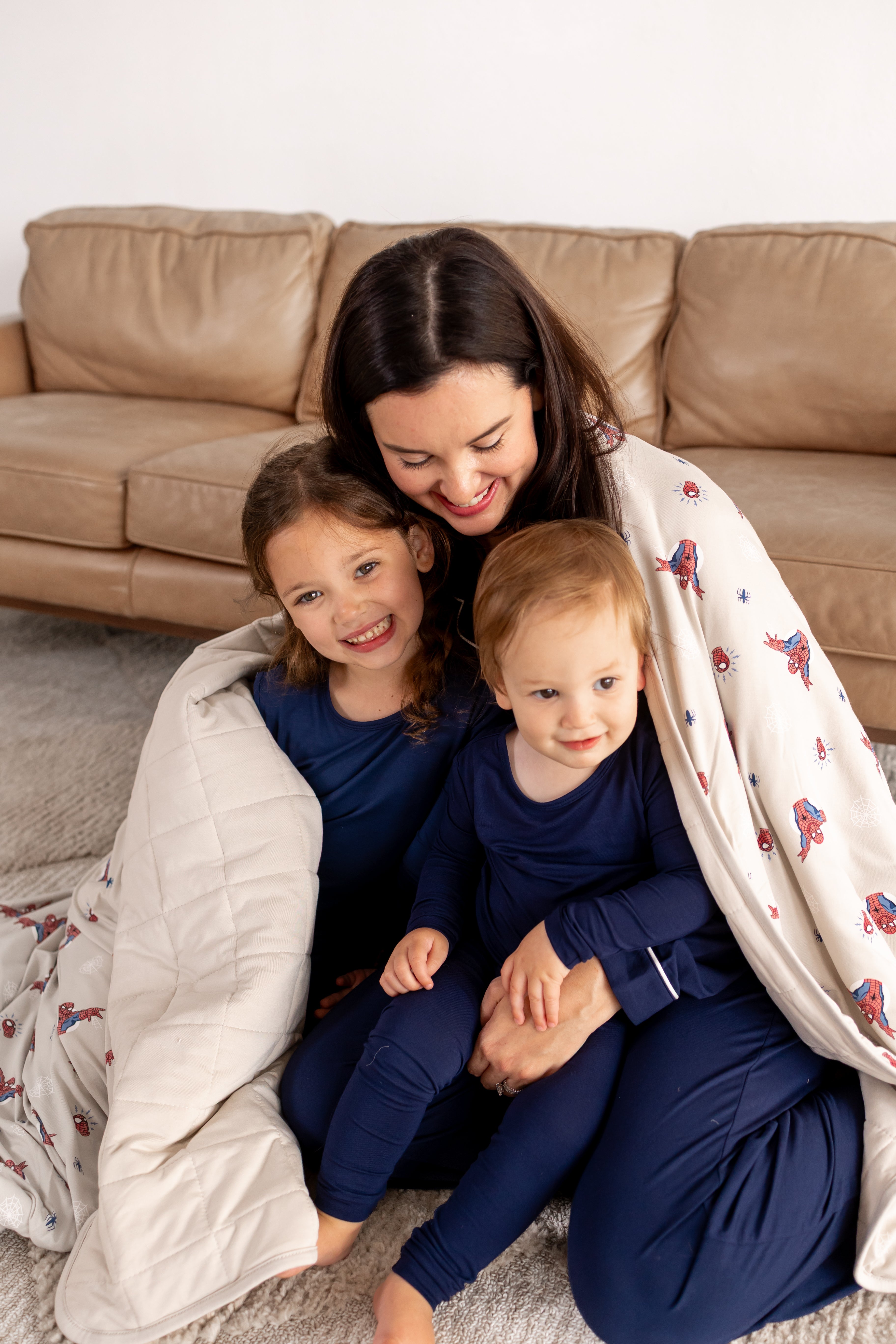 A woman and two kids posing in blue pajamas in front of beige couch wrapped in an oat color Adult Quilted Blanket with Spider-Man print