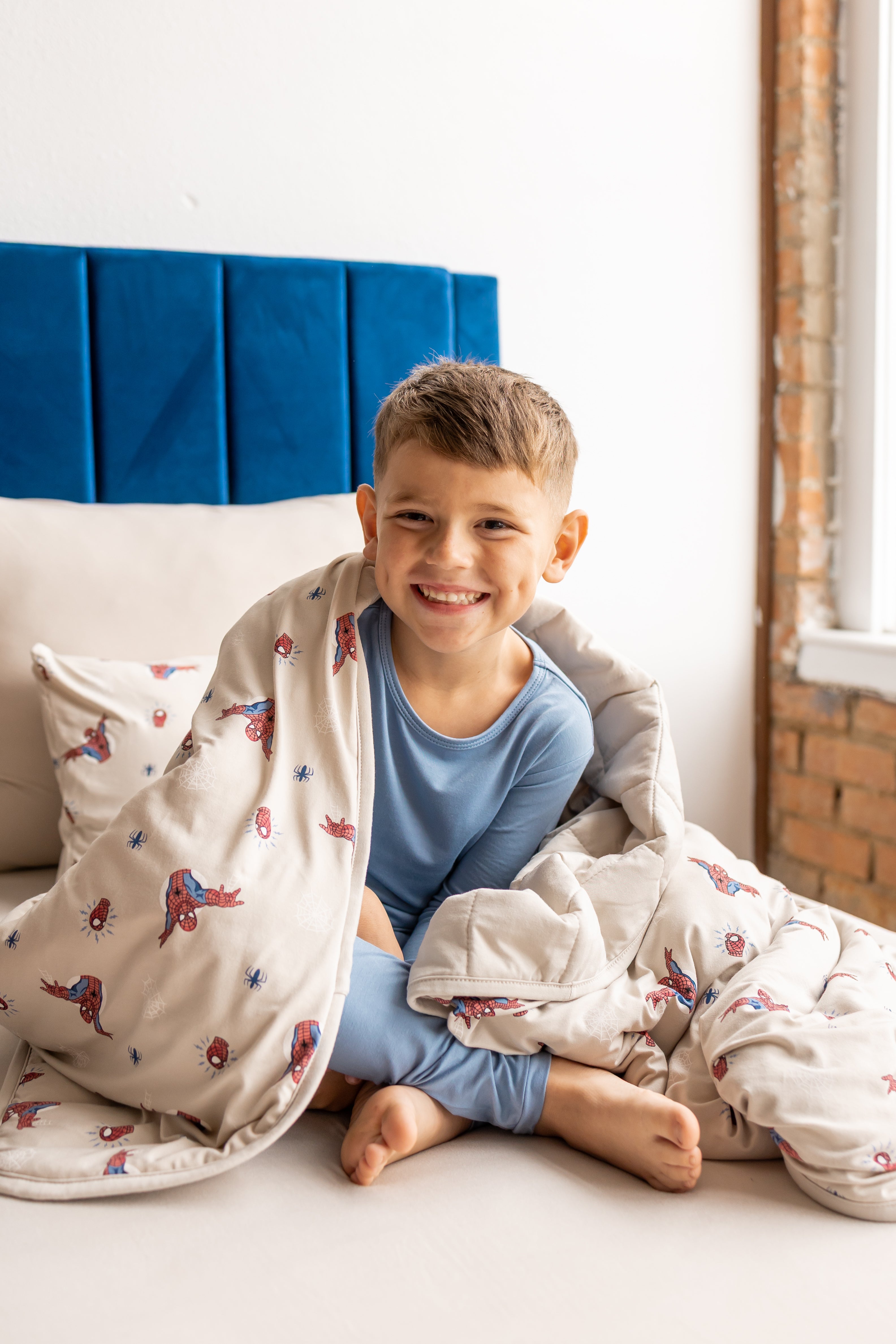 Boy sitting on a bed with oat color sheets and pillow wrapped in oat color Youth Blanket with Spider-Man print wearing blue pajamas