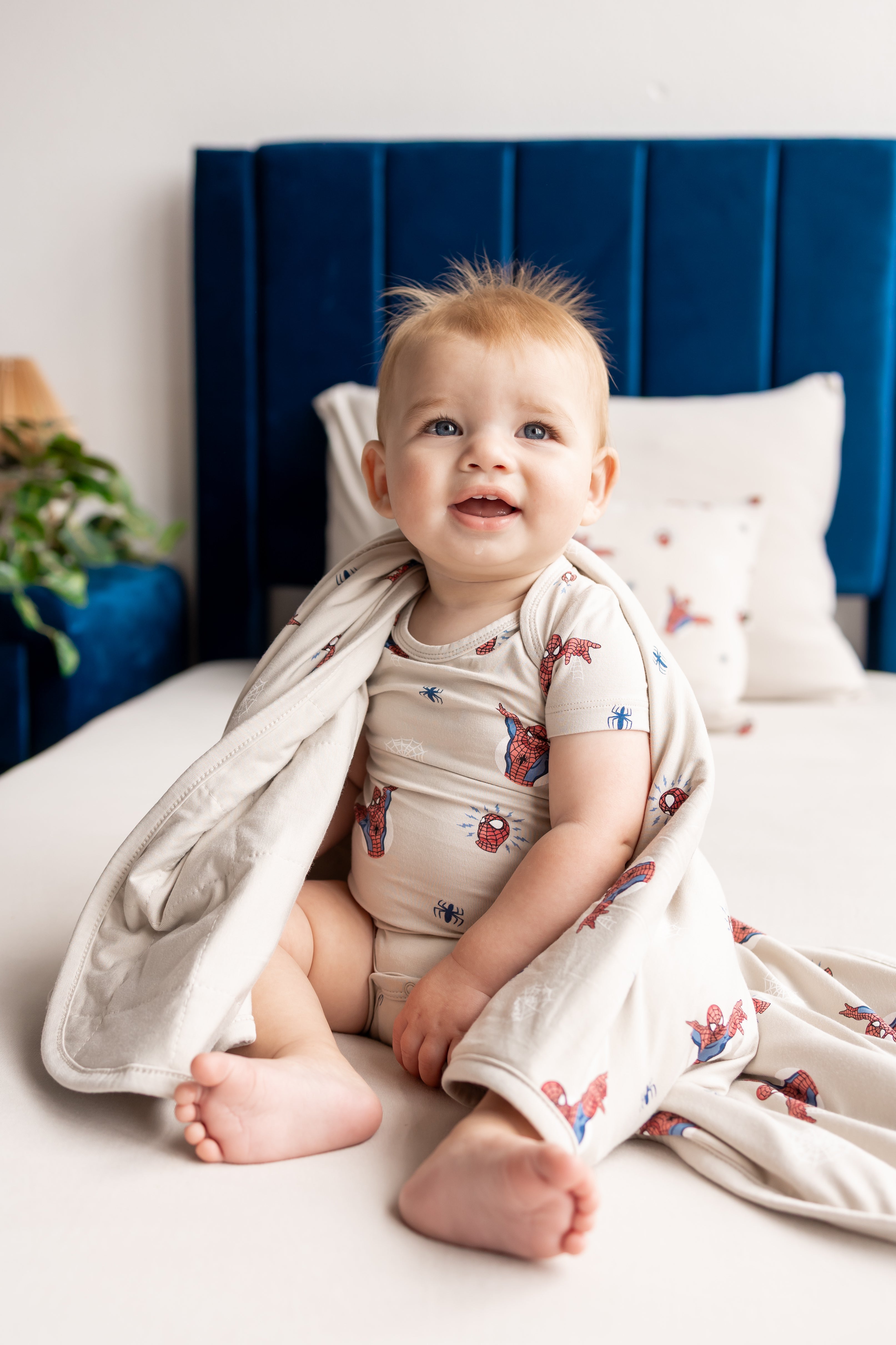 Baby sitting on a beige bed wrapped in an oat color Baby Blanket with Spider-Man print