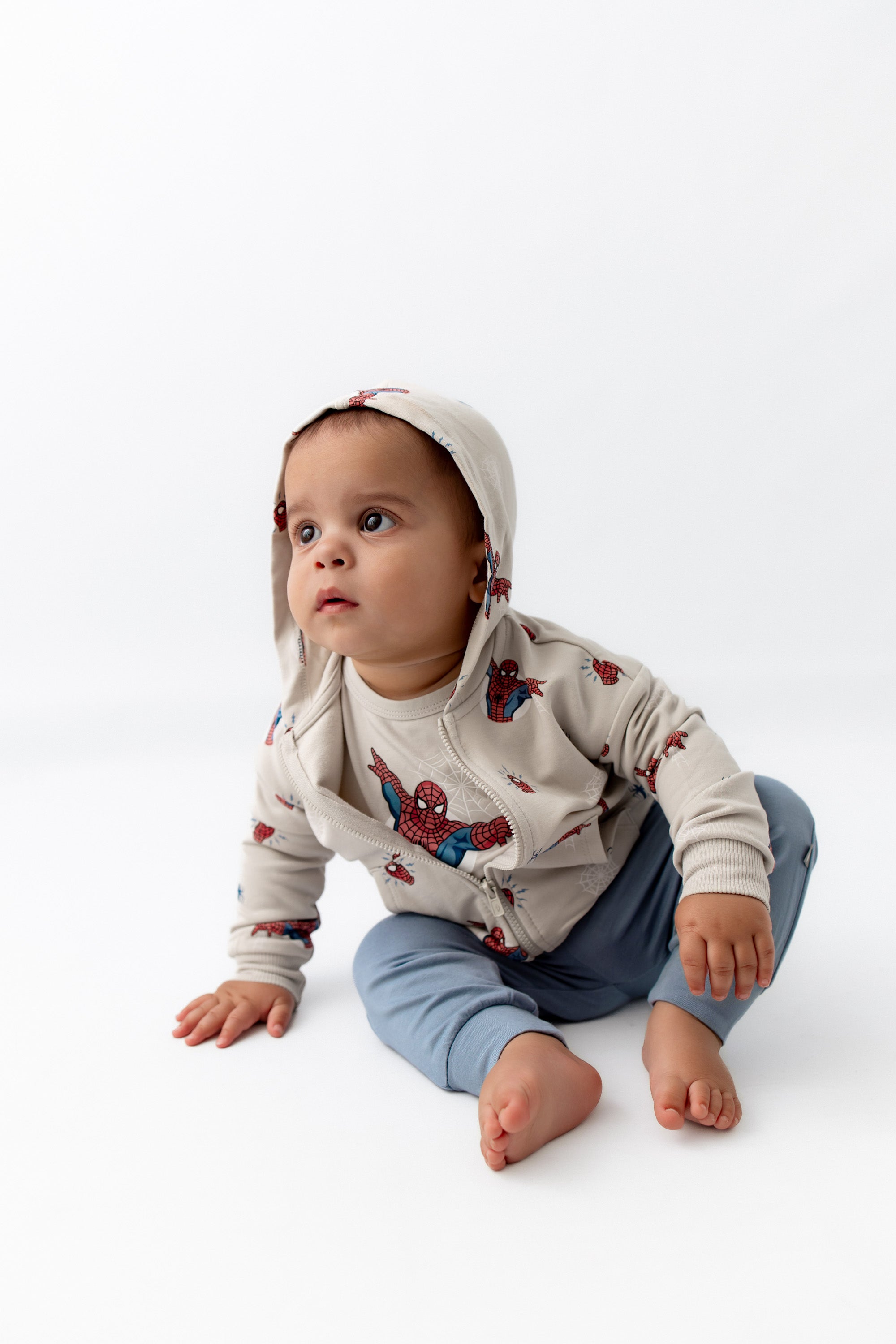 Toddler sitting down leaning over wearing a spiderman jacket with the hoodie on is head.