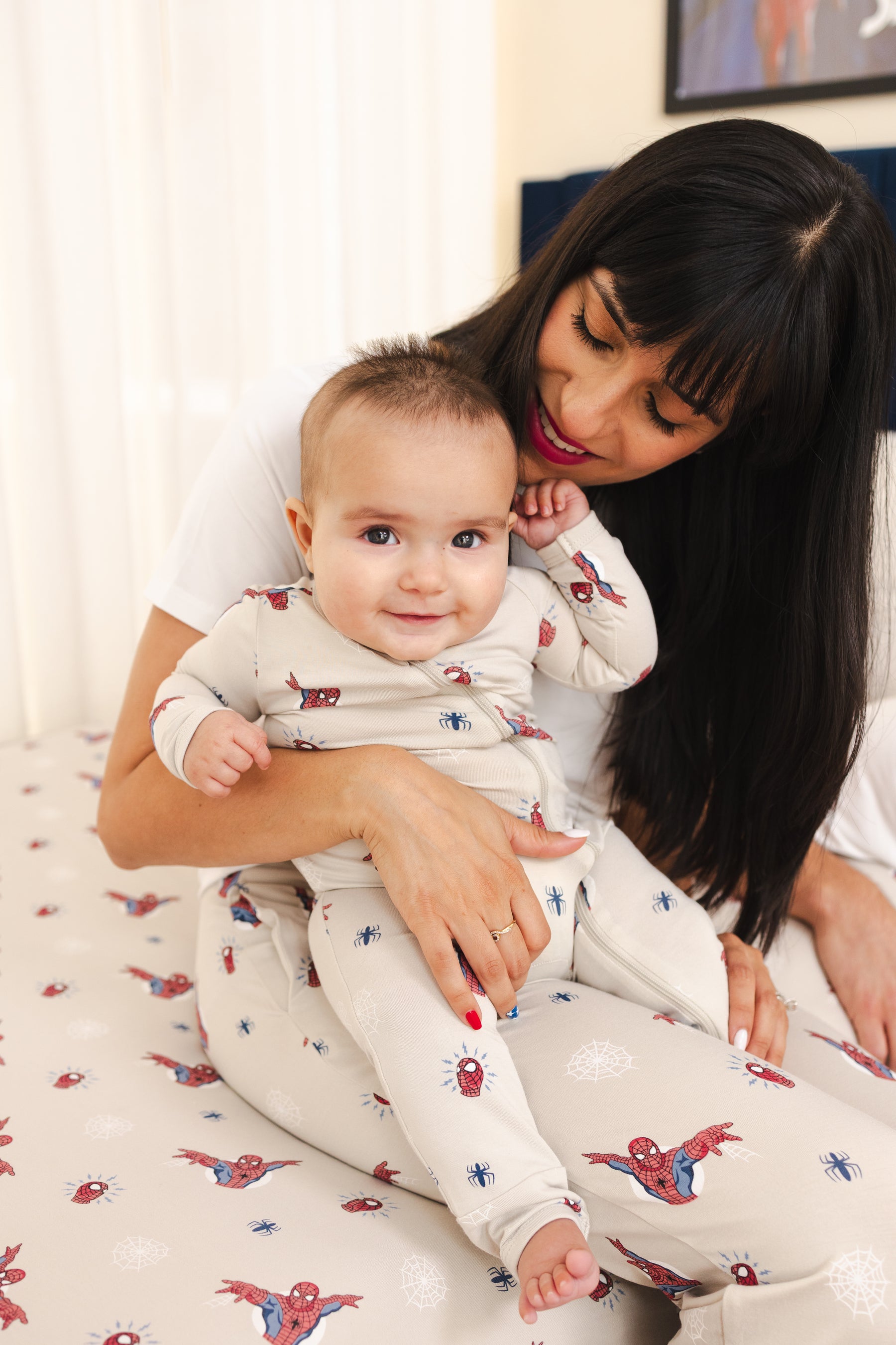 A baby and a woman wearing matching Spider-Man loungewear sitting on a bed. The pajamas are off-white with a small red and blue Spider-Man print.