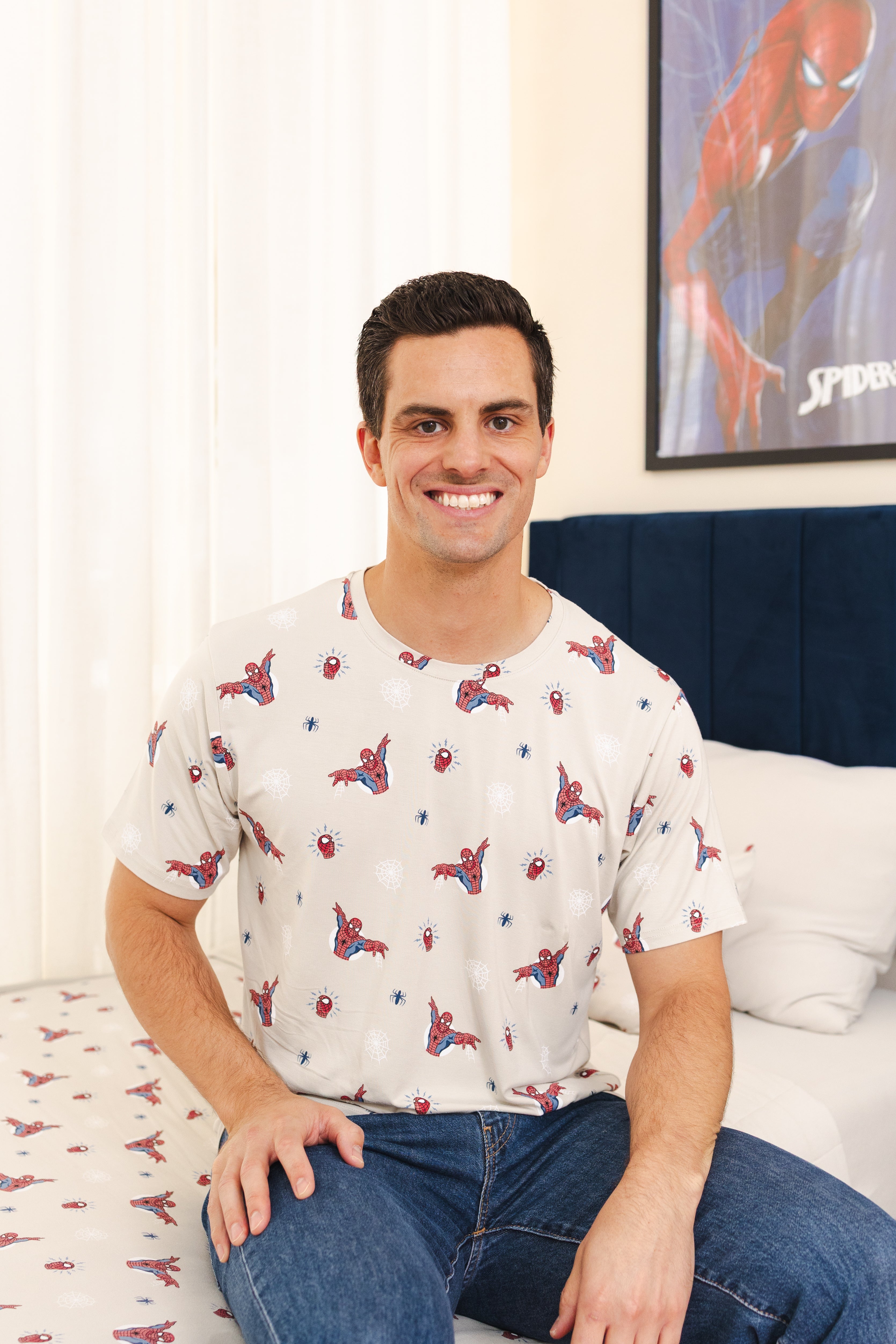 A man sitting on a bed wearing a white short-sleeve t-shirt with a red and blue Spider-Man print, paired with blue jeans. A Spider-Man poster is visible on the wall behind him.
