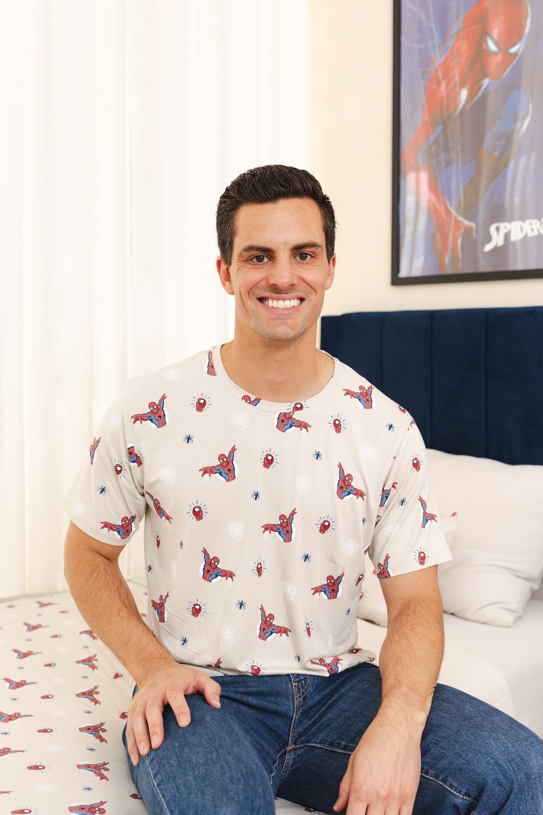 A man sitting on a bed wearing a white short-sleeve t-shirt with a red and blue Spider-Man print, paired with blue jeans. A Spider-Man poster is visible on the wall behind him.