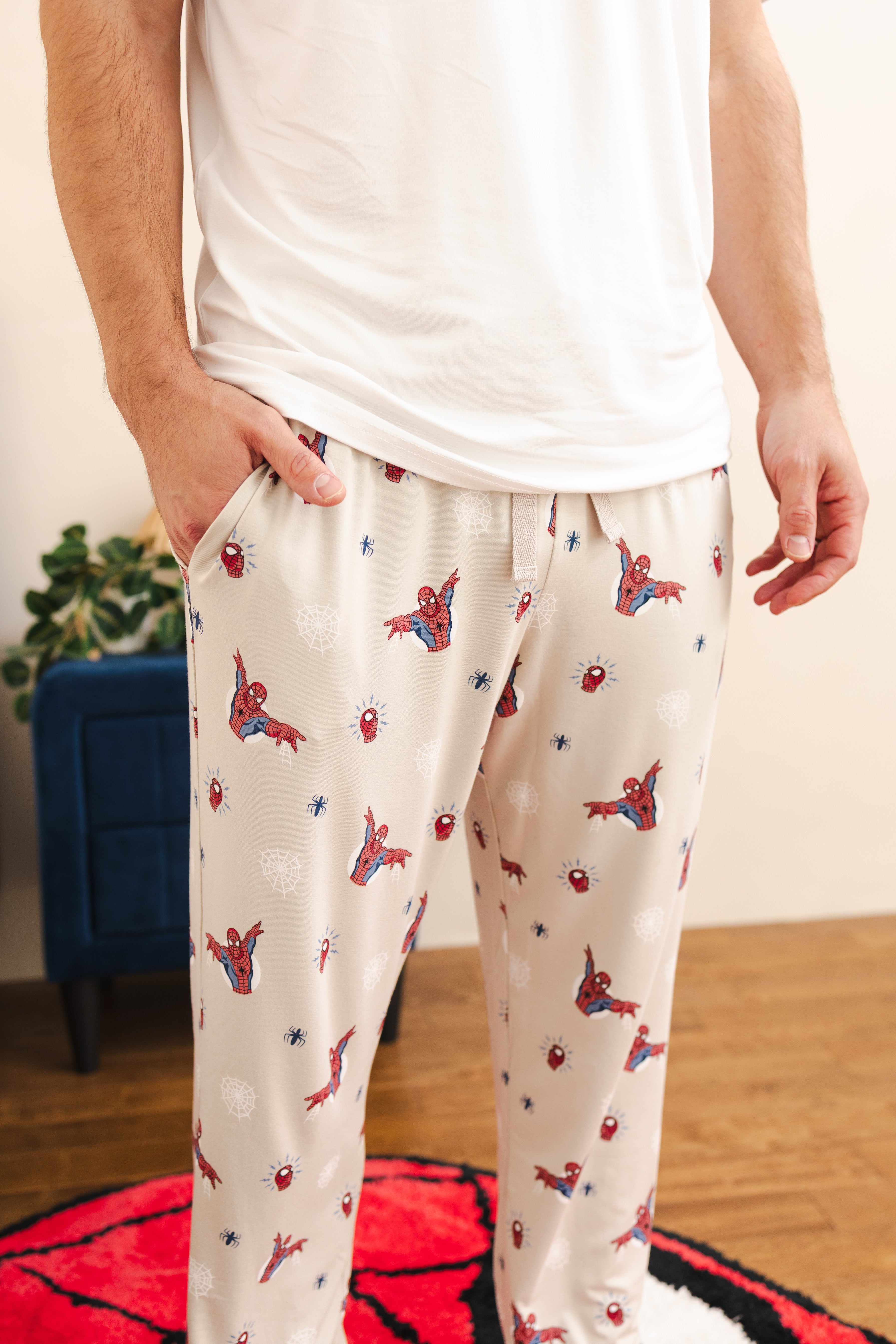 A person wearing cream-colored pajama pants with an all-over Spider-Man print, paired with a white t-shirt. The pants feature a drawstring waist and side pockets, shown in a home setting with a wooden floor and a blue cabinet in the background.