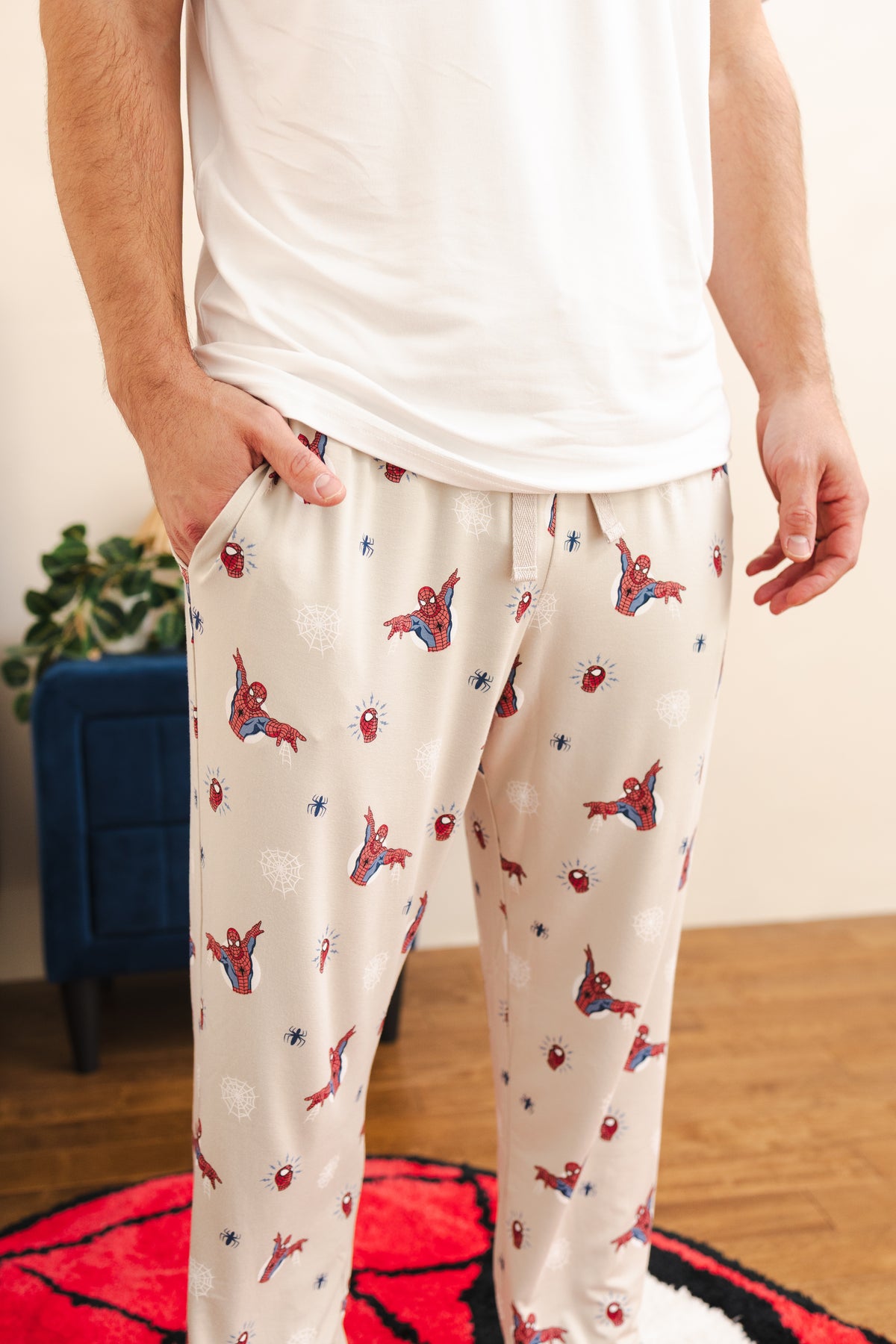 A person wearing cream-colored pajama pants with an all-over Spider-Man print, paired with a white t-shirt. The pants feature a drawstring waist and side pockets, shown in a home setting with a wooden floor and a blue cabinet in the background.