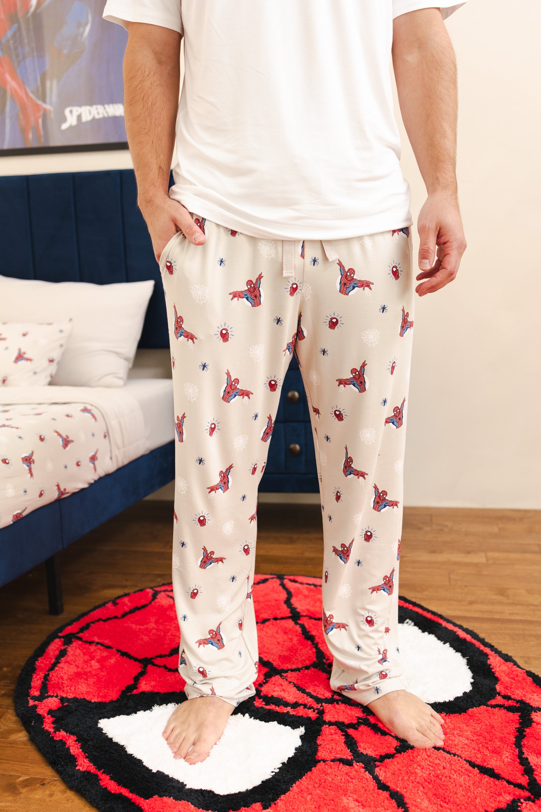 A person wearing cream-colored pajama pants with a red and blue Spider-Man spider and web pattern, standing barefoot on a red and black Spider-Man themed rug in a bedroom setting.