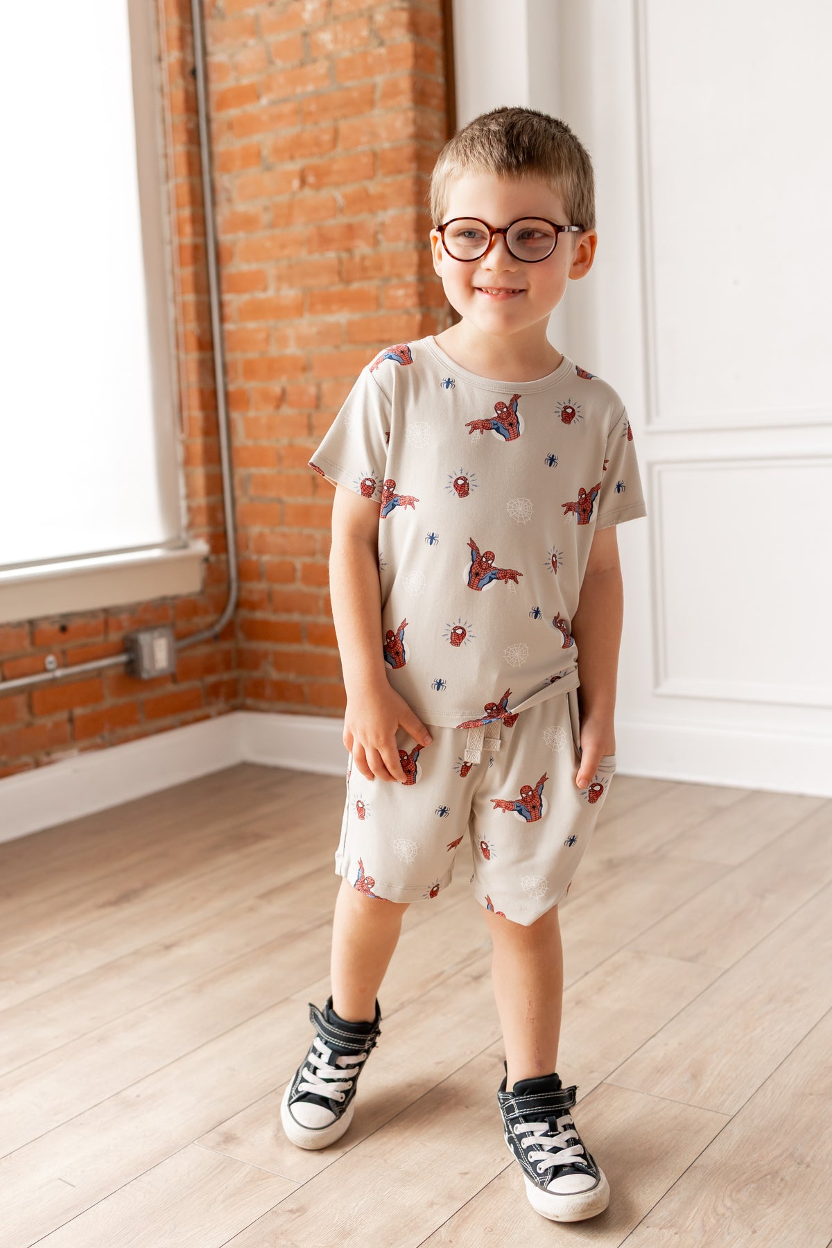 Boy with specs wearing oat color Toddler Basic Tee and Toddler Drawstring shorts with Spider-Man print in front of a brick wall and door